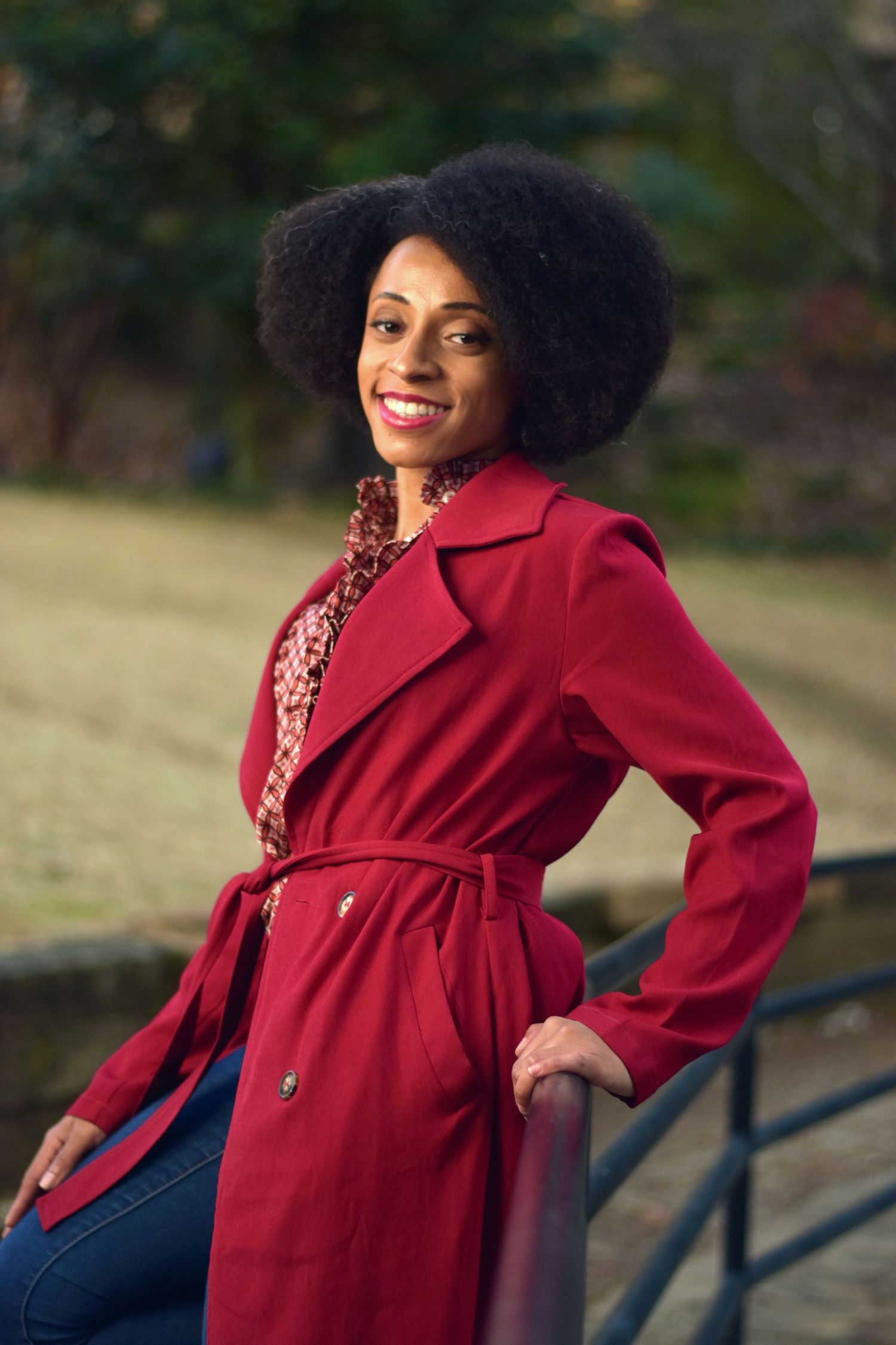 Smiling Black woman with afro hair poses in red trench coat outdoors in park setting.