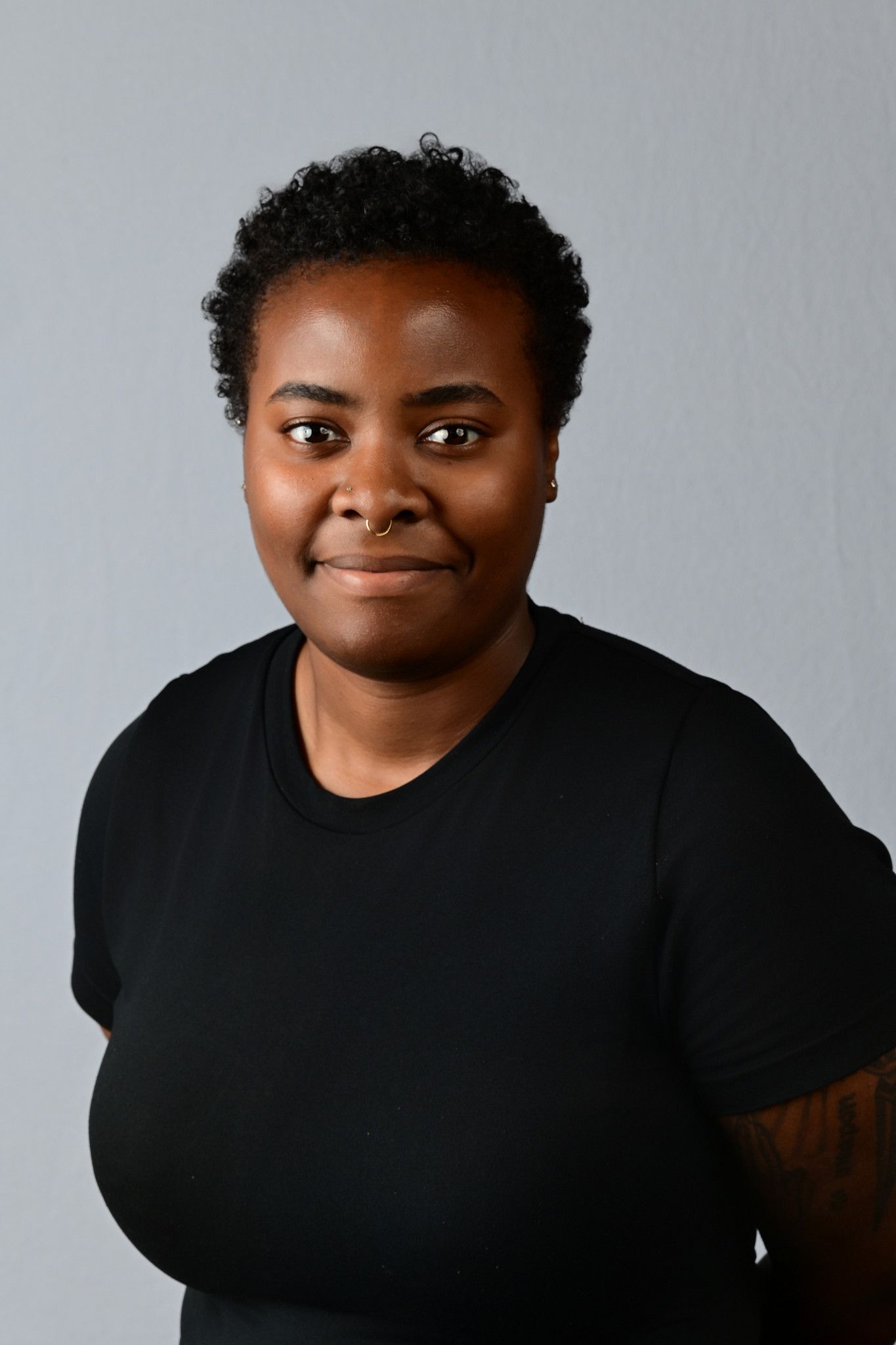 Young Black woman with short curly hair wearing a black t-shirt, smiling confidently against a gray background.