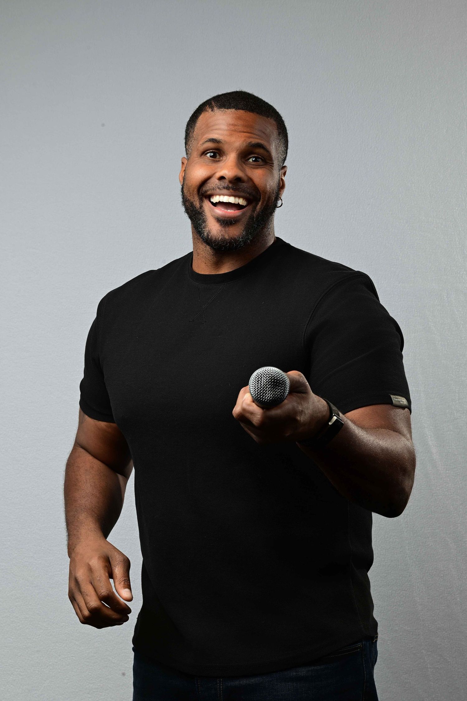 Smiling Black man in black t-shirt holding a microphone, performing stand-up comedy against a gray background.