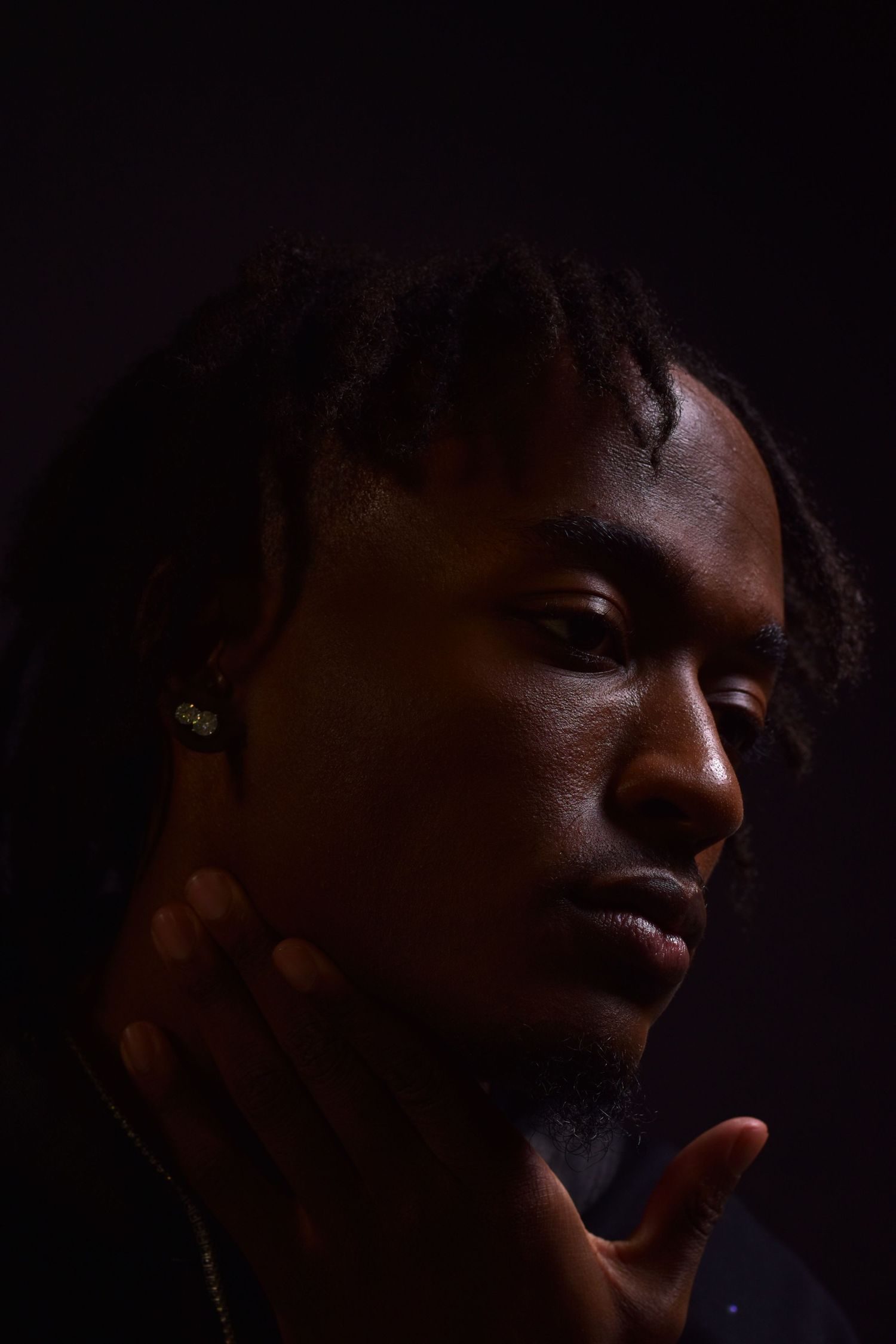 Close-up portrait of young Black man with dreadlocks posing thoughtfully against dark background.