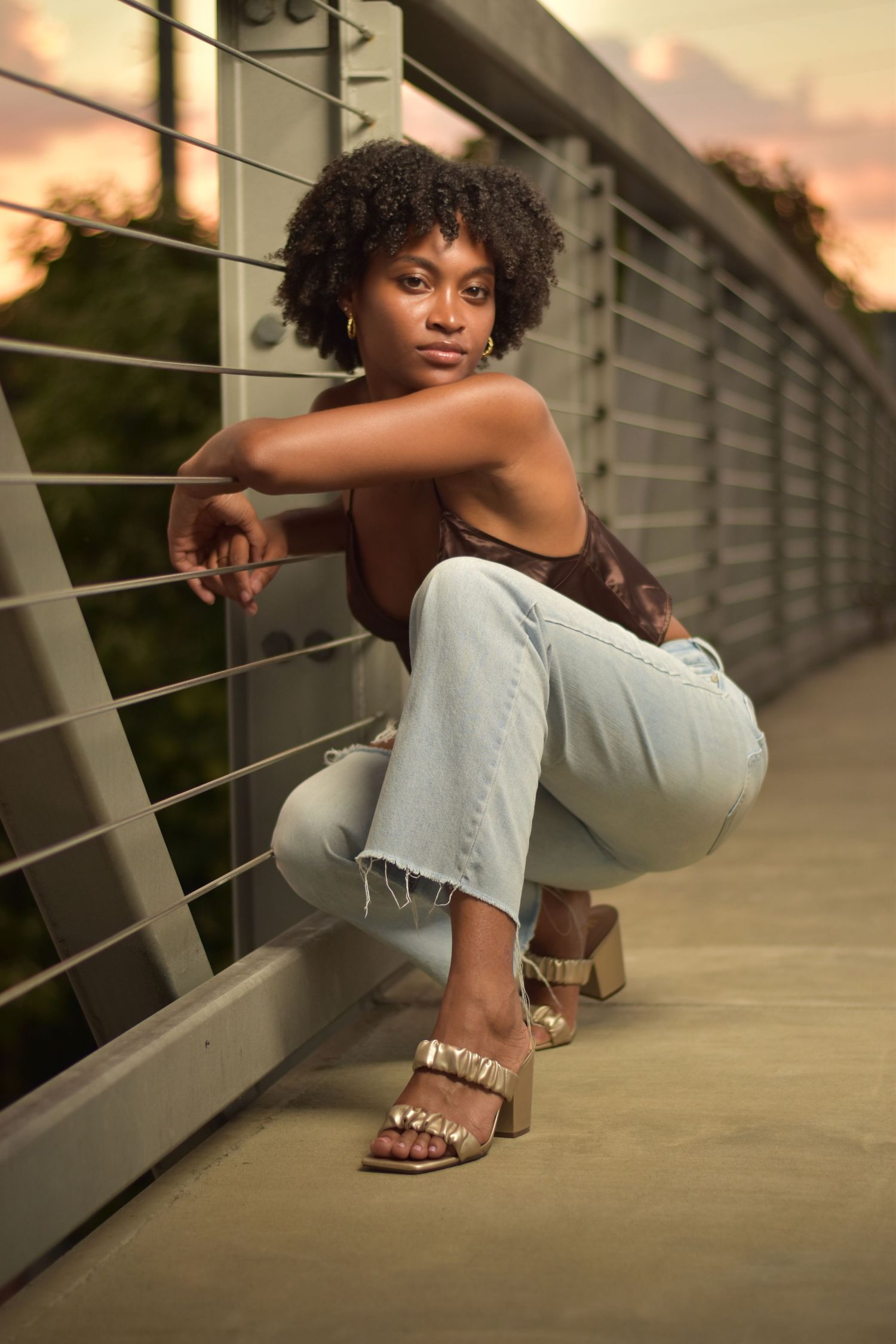 Young Black woman in jeans and gold heels crouching by metal railing on bridge at sunset.