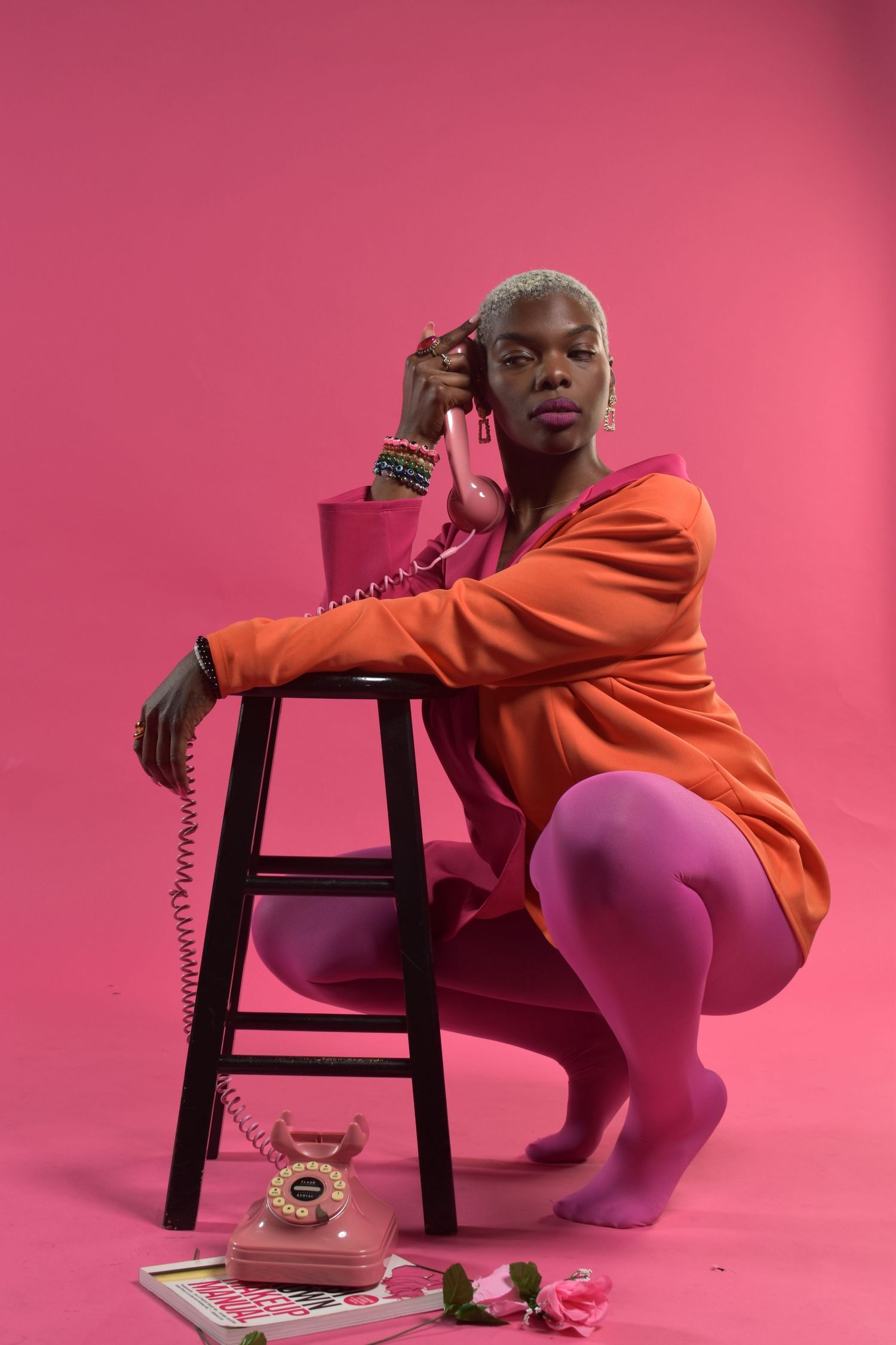 A Black woman in an orange dress and pink tights crouches by a stool, holding a pink vintage telephone receiver.