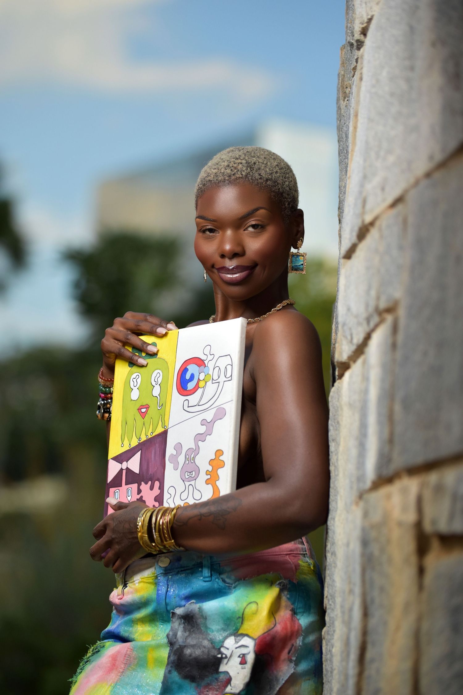 Smiling Black woman holding colorful illustrated canvas art, leaning against stone wall in tie-dye outfit outdoors.