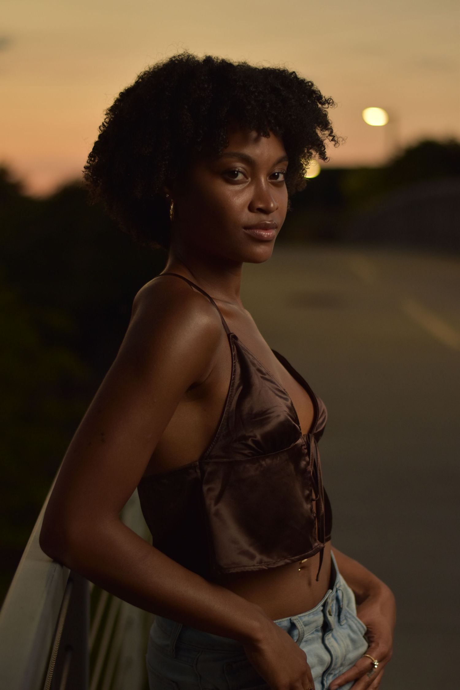 Black woman in brown satin crop top posing on road during vibrant orange and pink sunset.