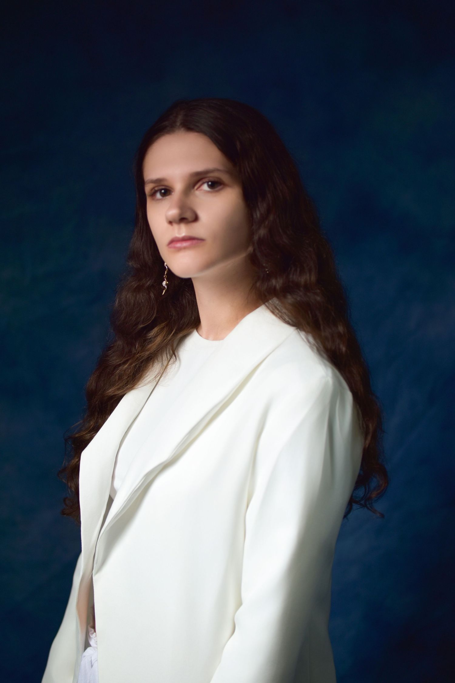 Young woman with long brown hair wearing a white blazer poses against a dark blue studio background.