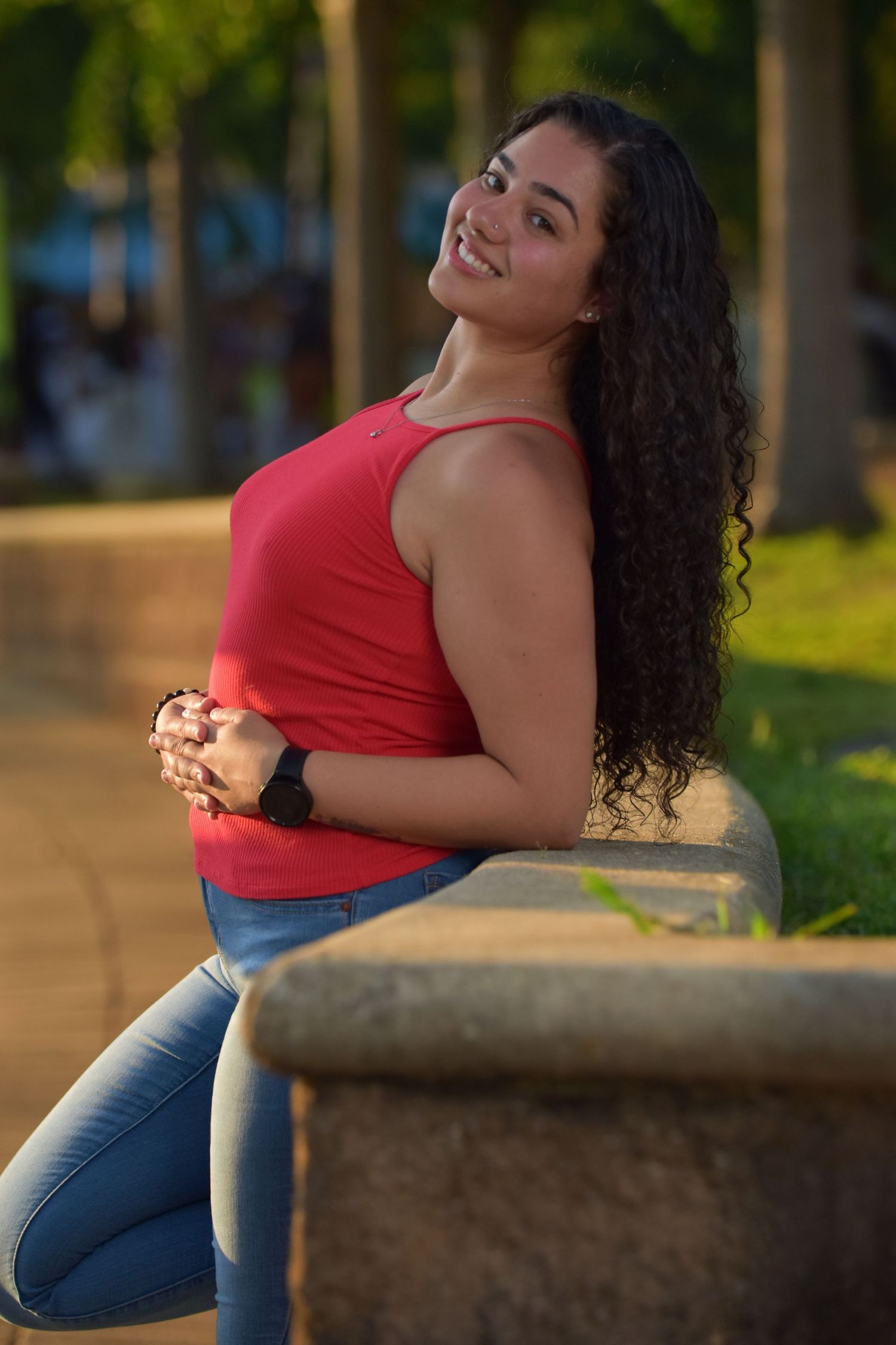 Smiling young woman with curly hair in red tank top leans on stone bench in sunny park.