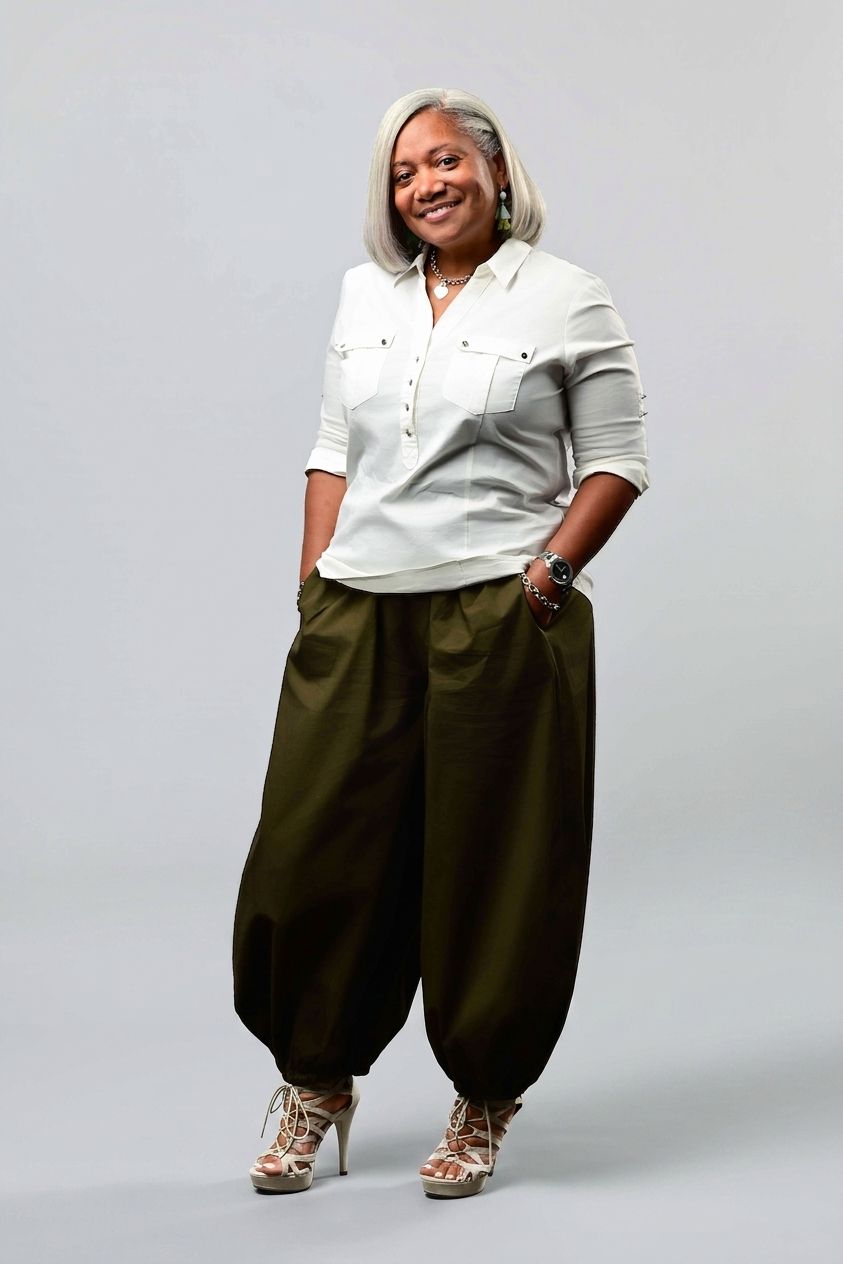 Smiling woman with silver hair wearing a white button-up shirt, olive green harem pants and strappy beige high heels, hands in pockets.