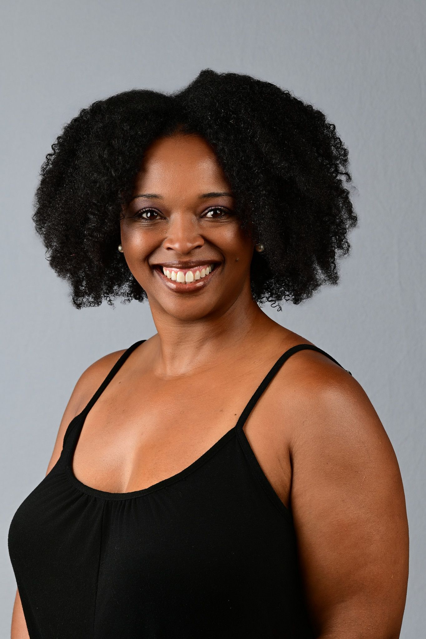 Smiling Black woman with natural afro hair wearing a black tank top against a gray background.