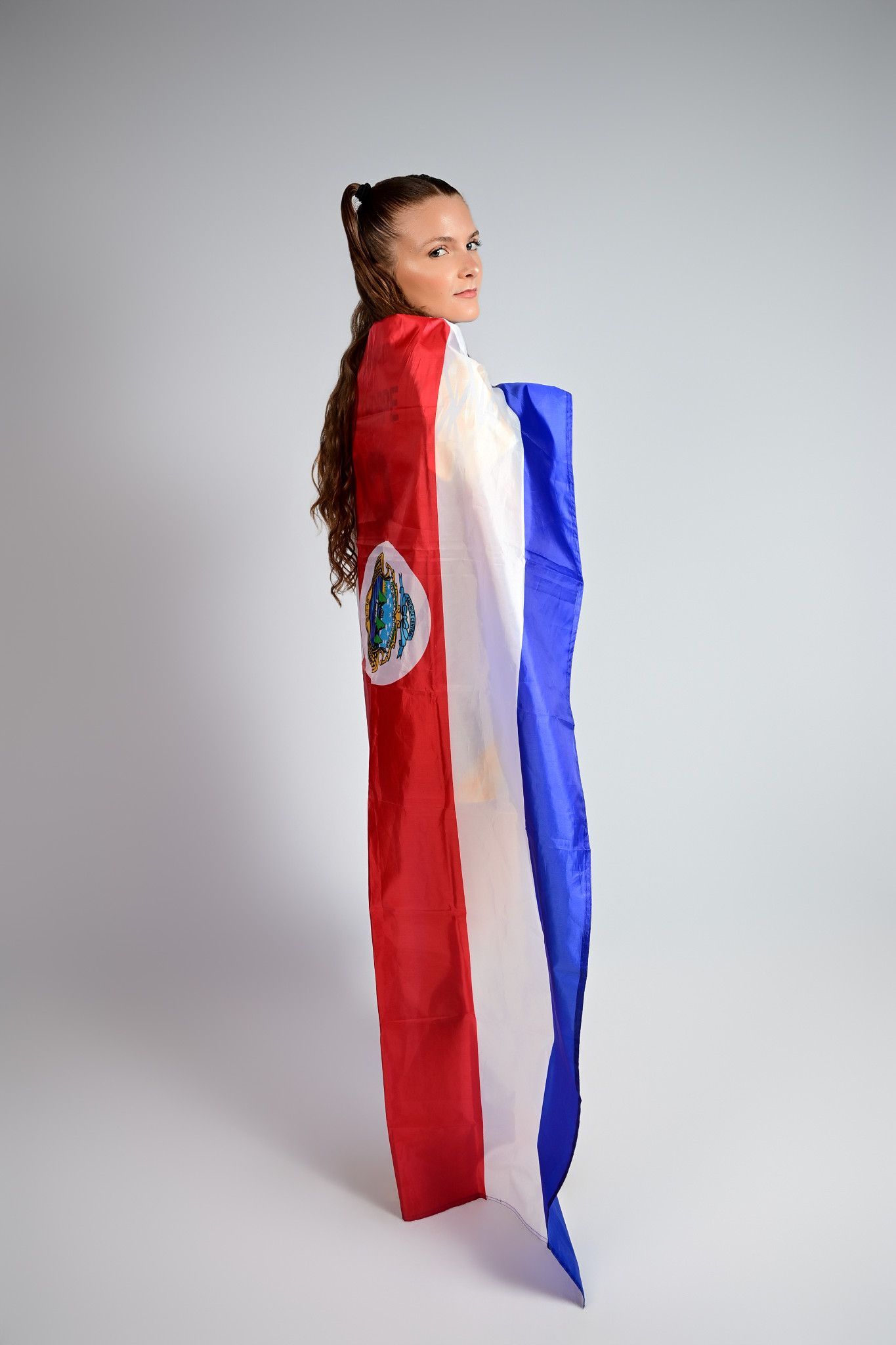 Young woman wrapped in Costa Rica flag, standing against gray background, looking over shoulder.