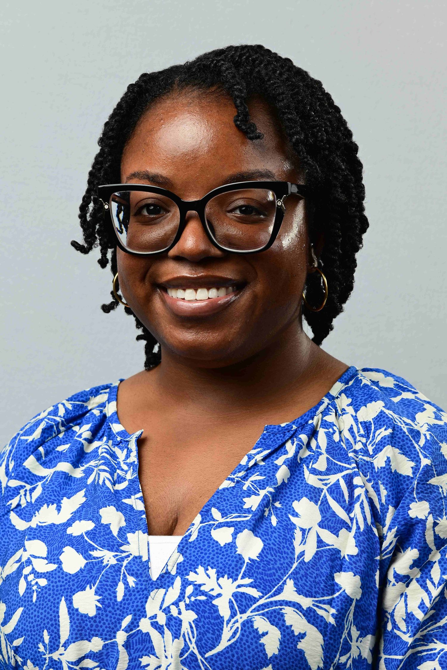 Smiling young Black woman wearing glasses and blue floral blouse poses for a professional headshot portrait.