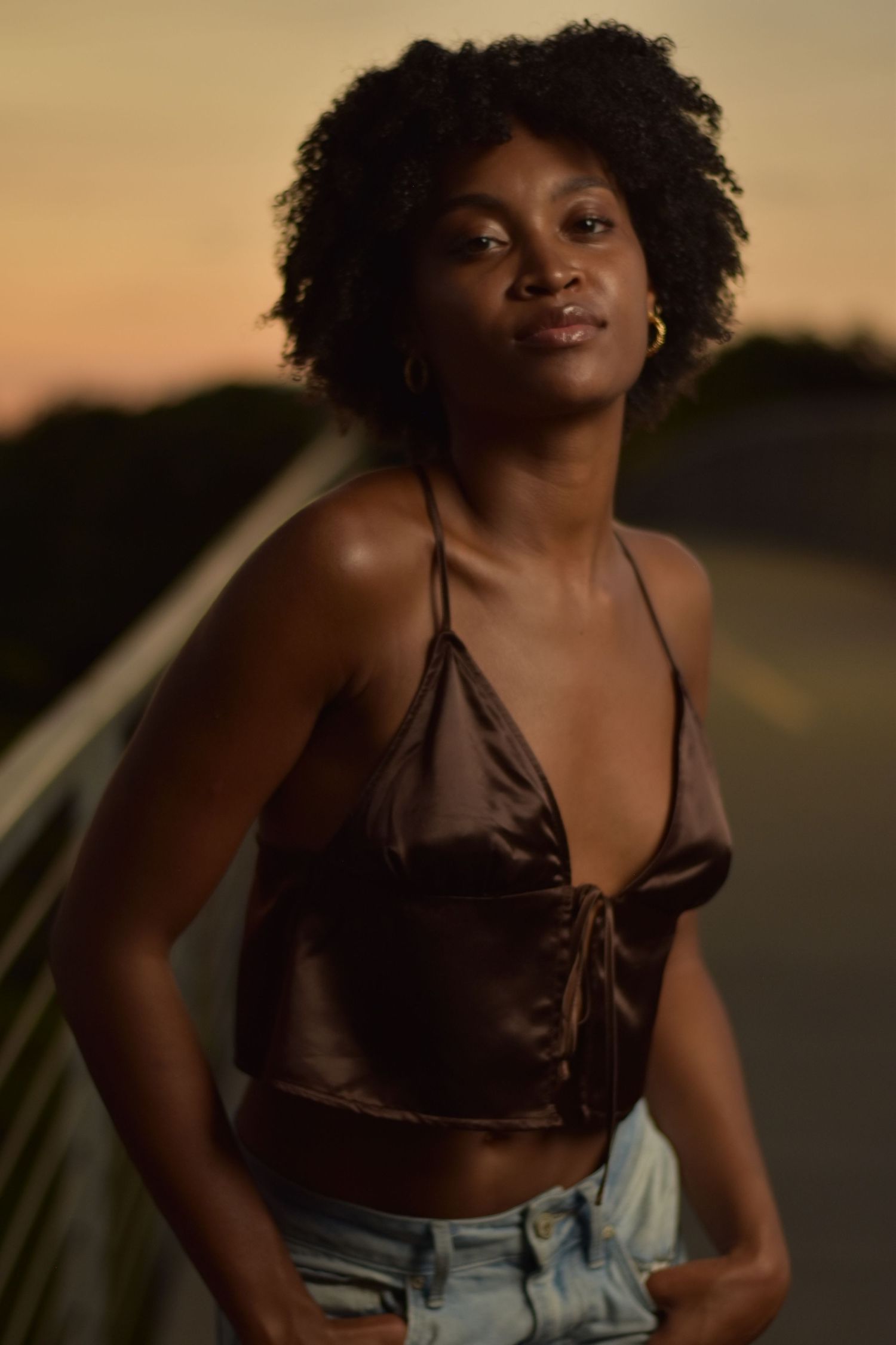 Young Black woman in brown halter top and light jeans smiling softly during warm golden sunset.