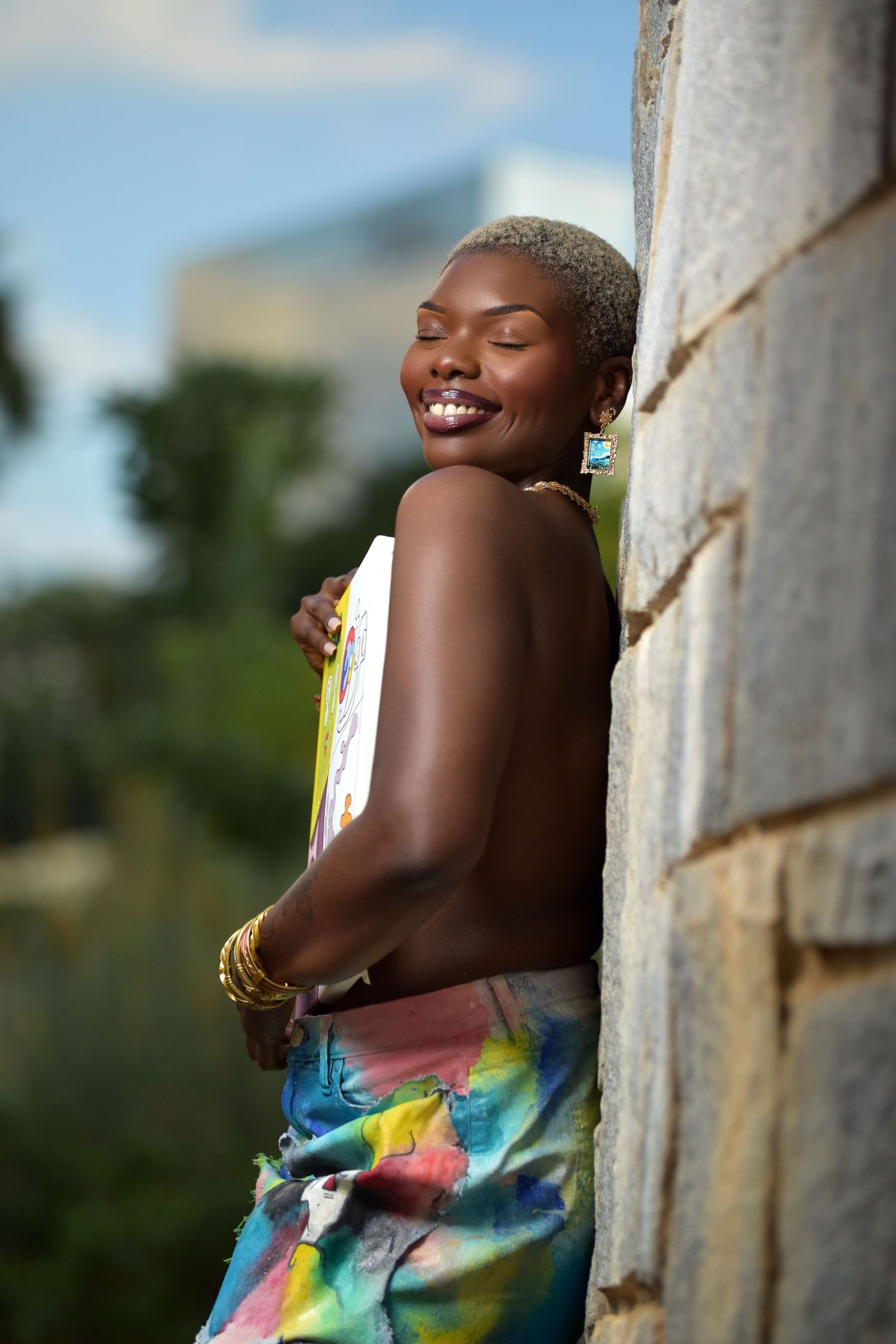 Joyful Black woman with eyes closed hugging colorful art canvas against stone wall, wearing tie-dye skirt and bangles.