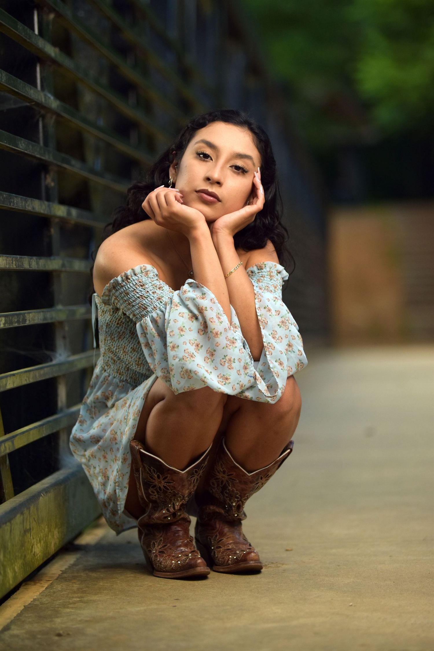 Young woman in floral off-shoulder dress and cowboy boots crouching against metal fence outdoors.