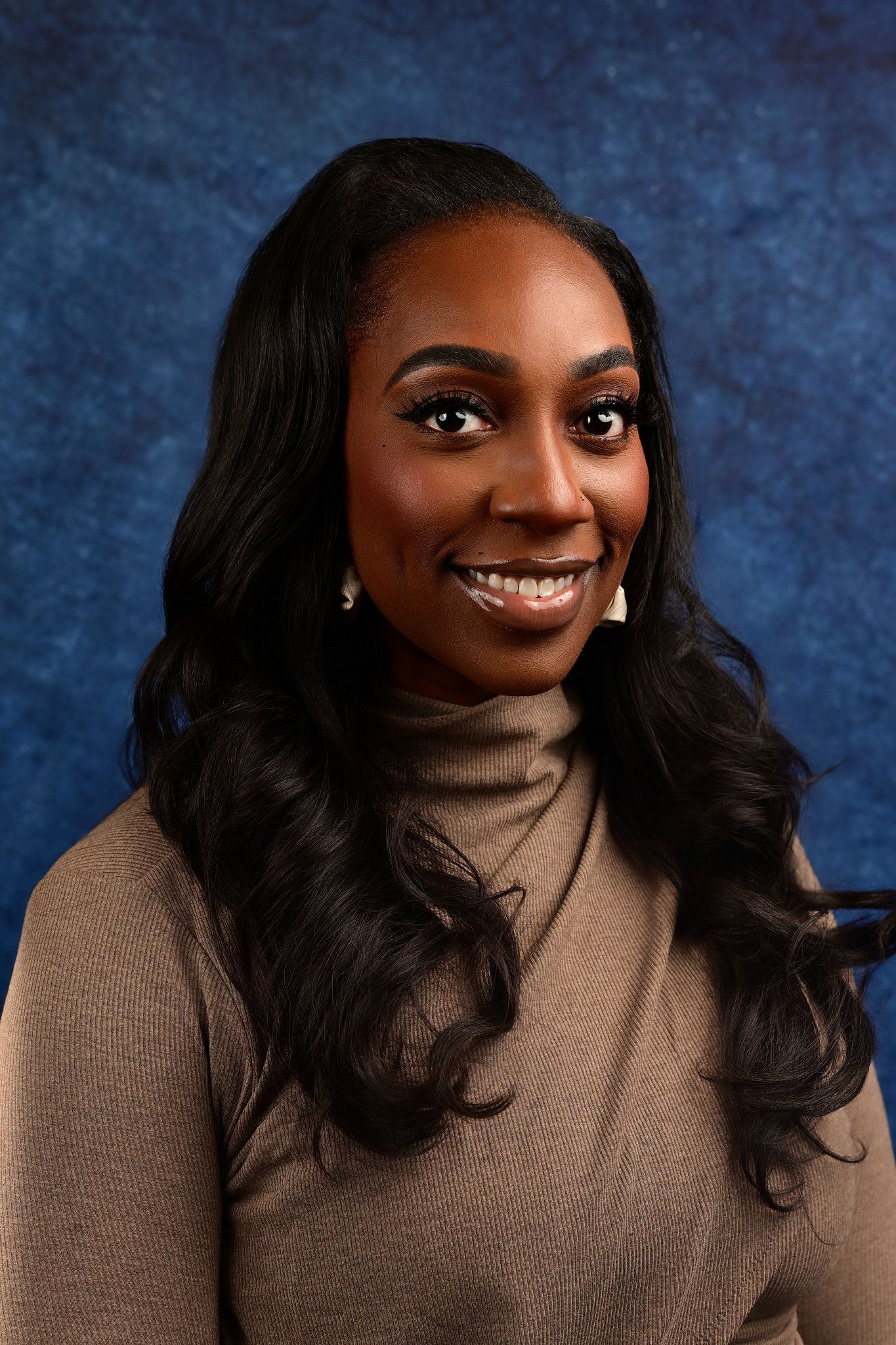 Smiling woman with long wavy black hair wears a tan turtleneck sweater in a studio portrait against a blue textured backdrop.