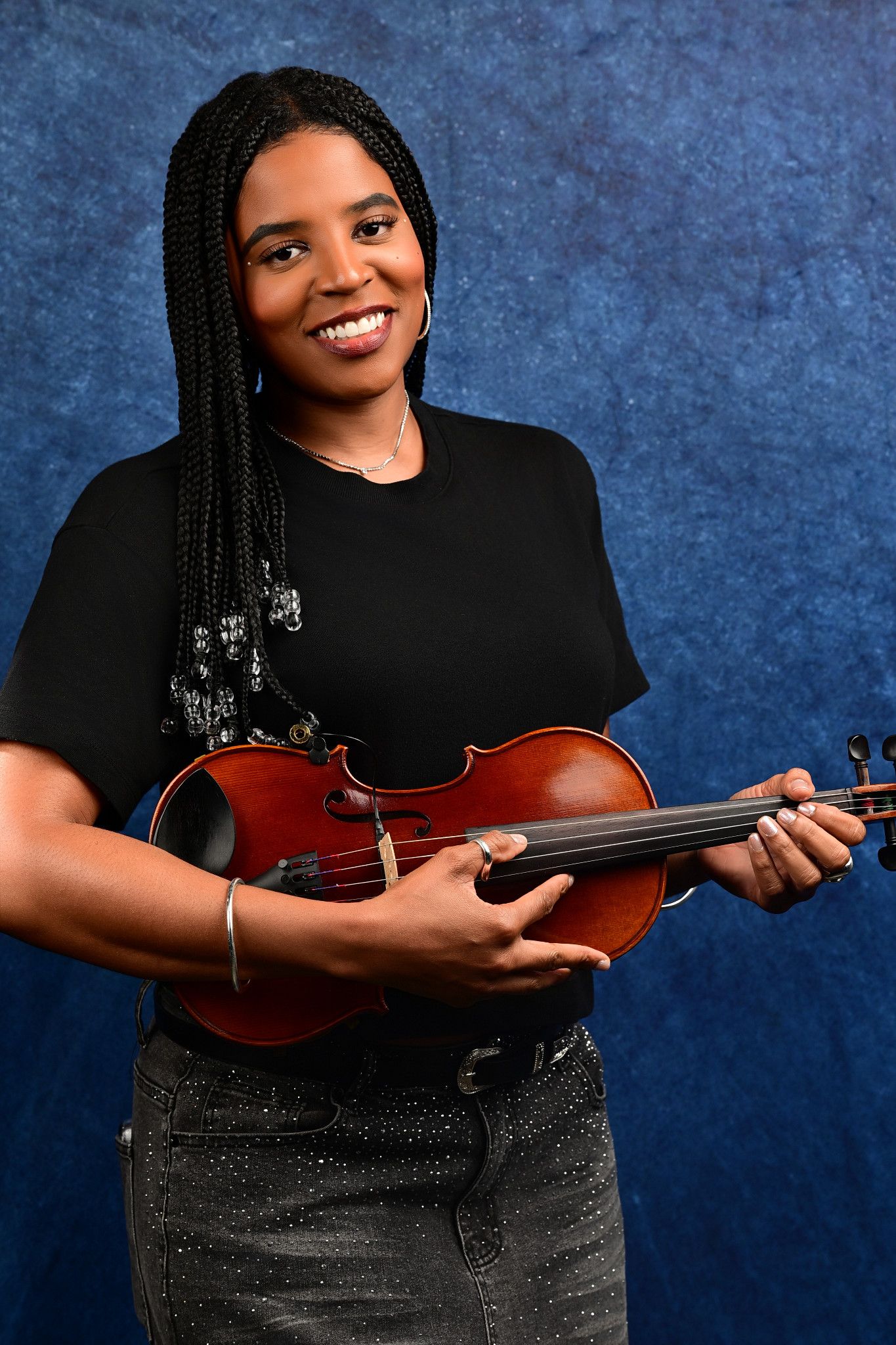 Smiling woman with long braids in a black t-shirt and sparkly jeans holds a violin against a blue textured studio backdrop.