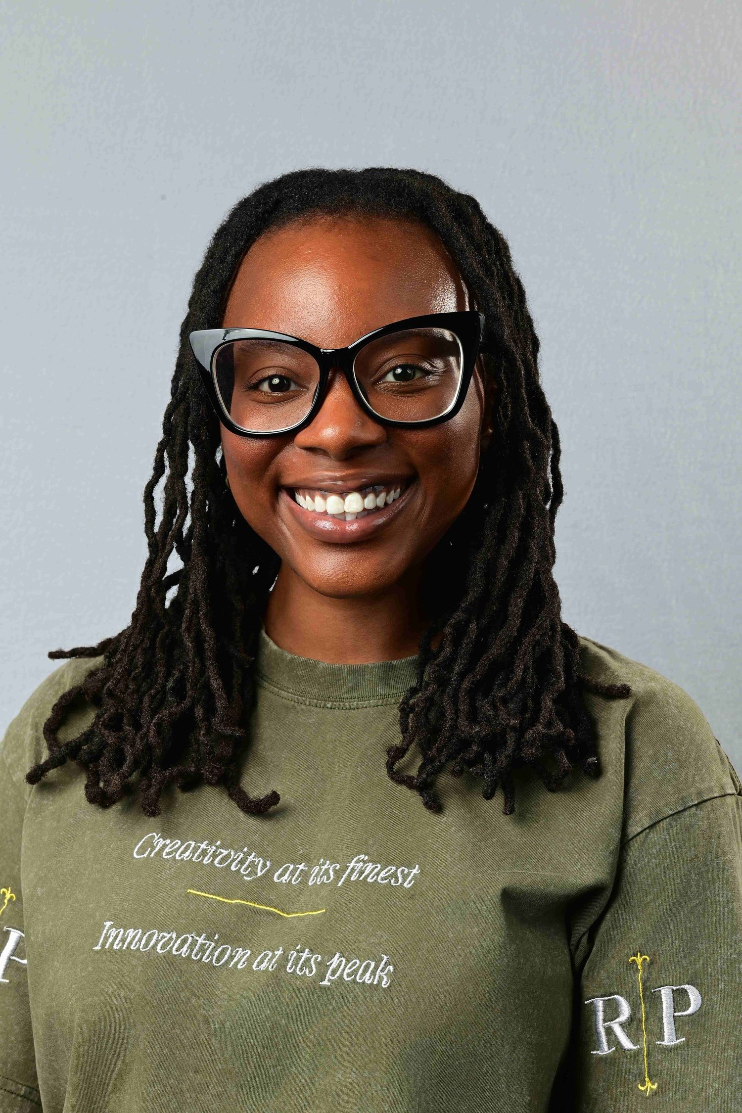 Smiling young woman with locs and black glasses wearing an olive green sweatshirt.