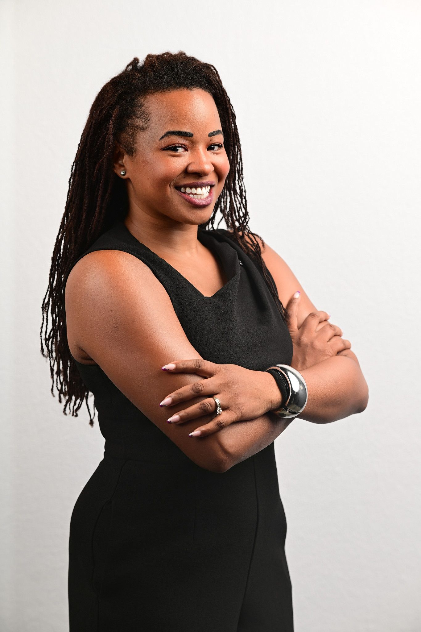 Smiling woman with long locs and arms crossed poses confidently in a sleeveless black outfit against a plain light background.