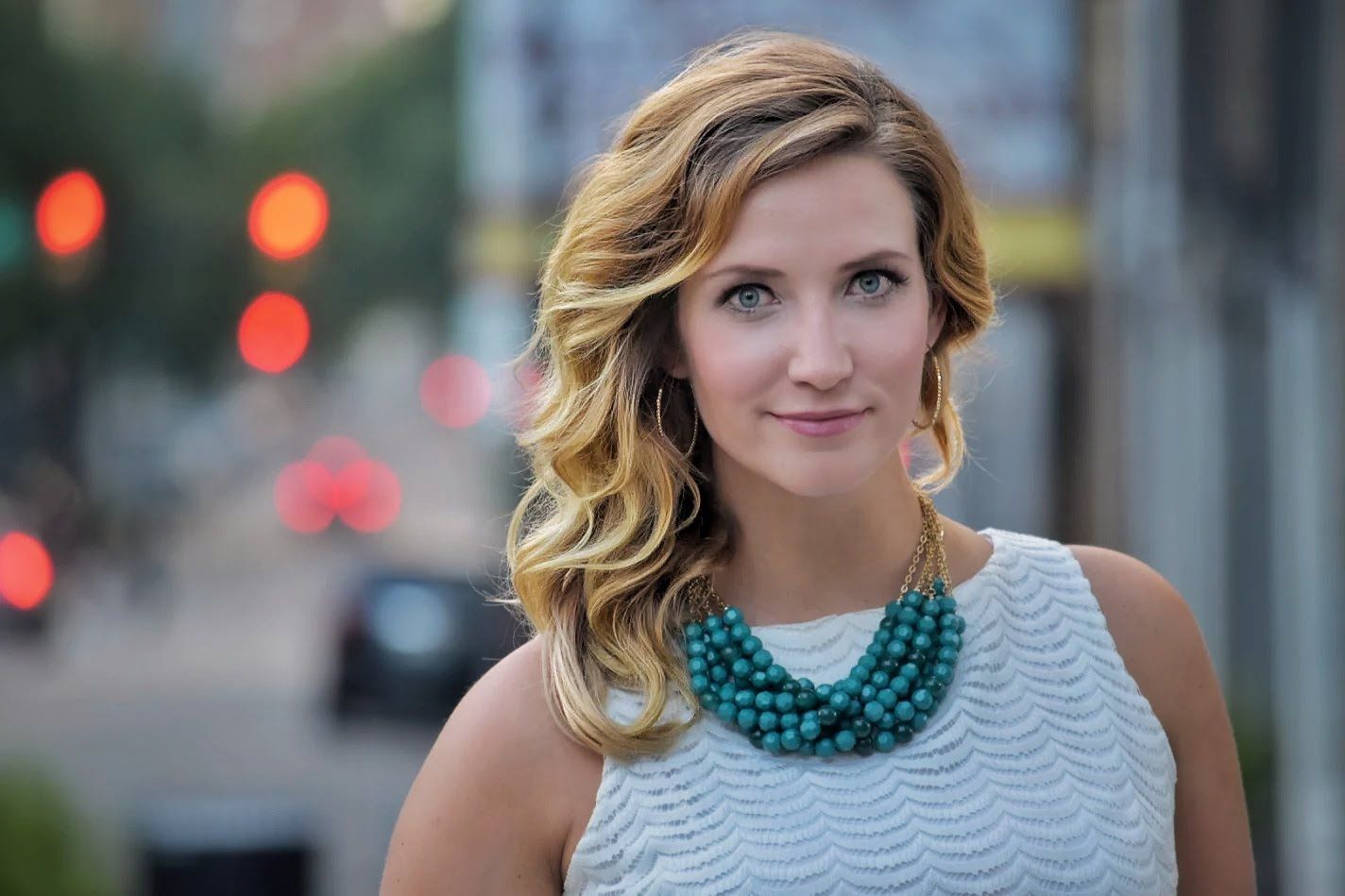 Woman with blonde wavy hair wearing a teal beaded necklace and white top, posing on an urban street.