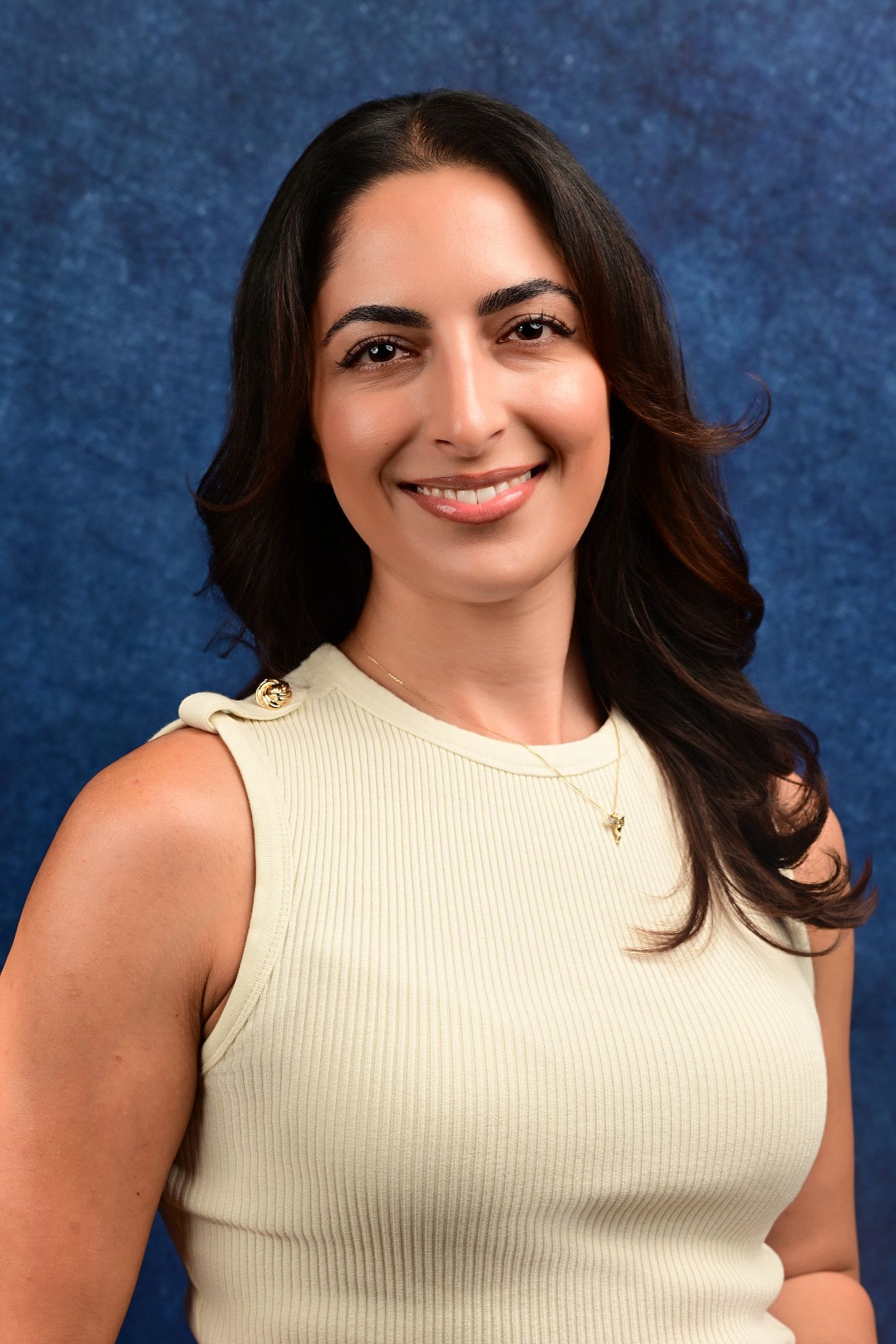 Smiling woman with long dark wavy hair wearing a cream sleeveless ribbed top and gold necklace against a blue textured backdrop.