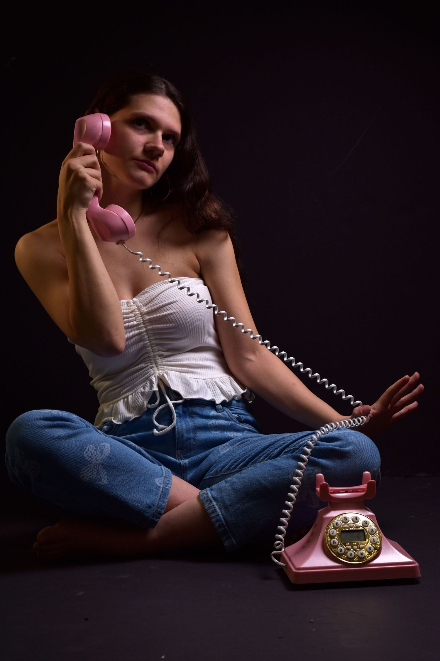 Young woman sits cross-legged holding vintage pink rotary telephone against dark studio background.