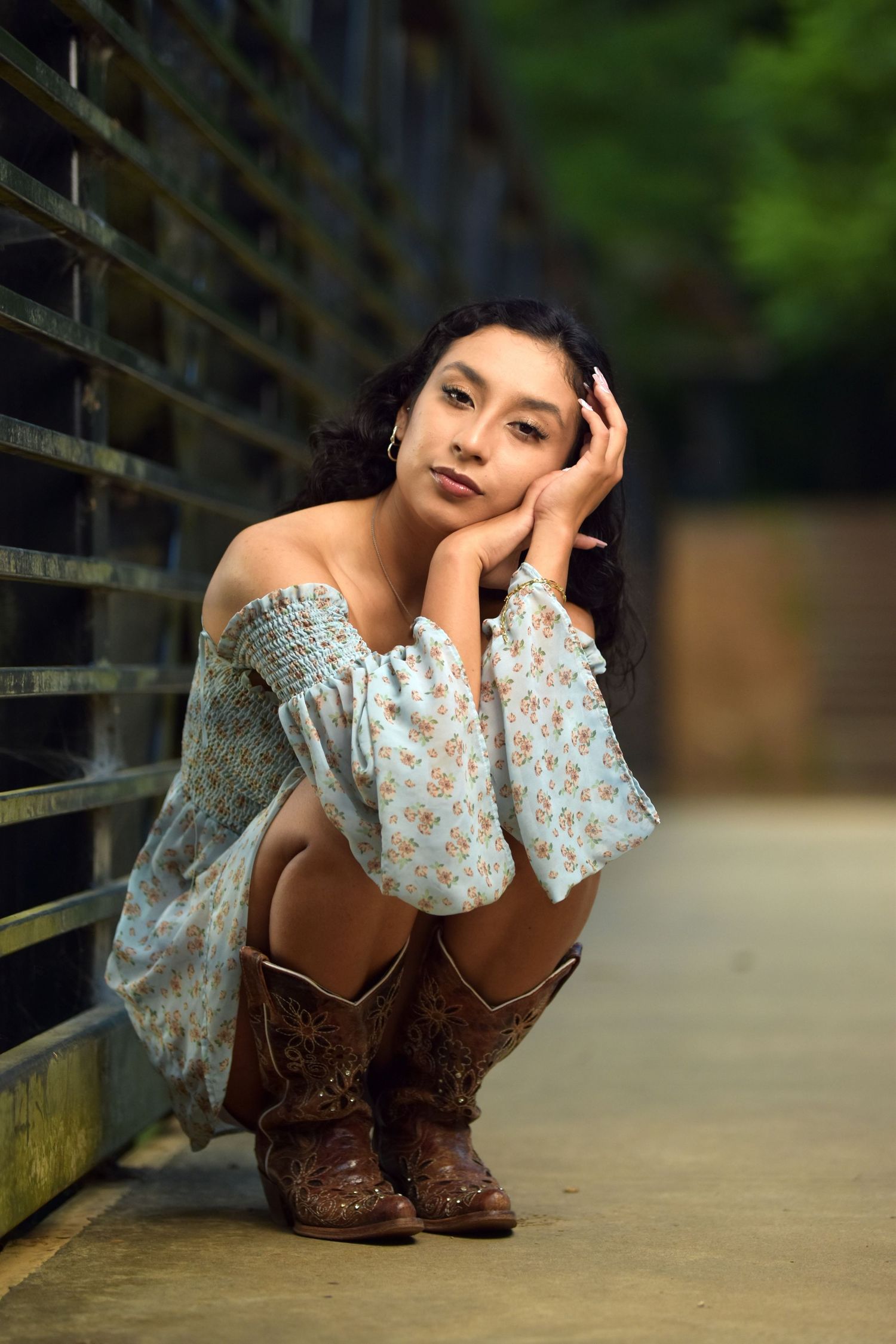 Young woman in light floral off-shoulder dress and cowboy boots sitting crouched near metal fence.