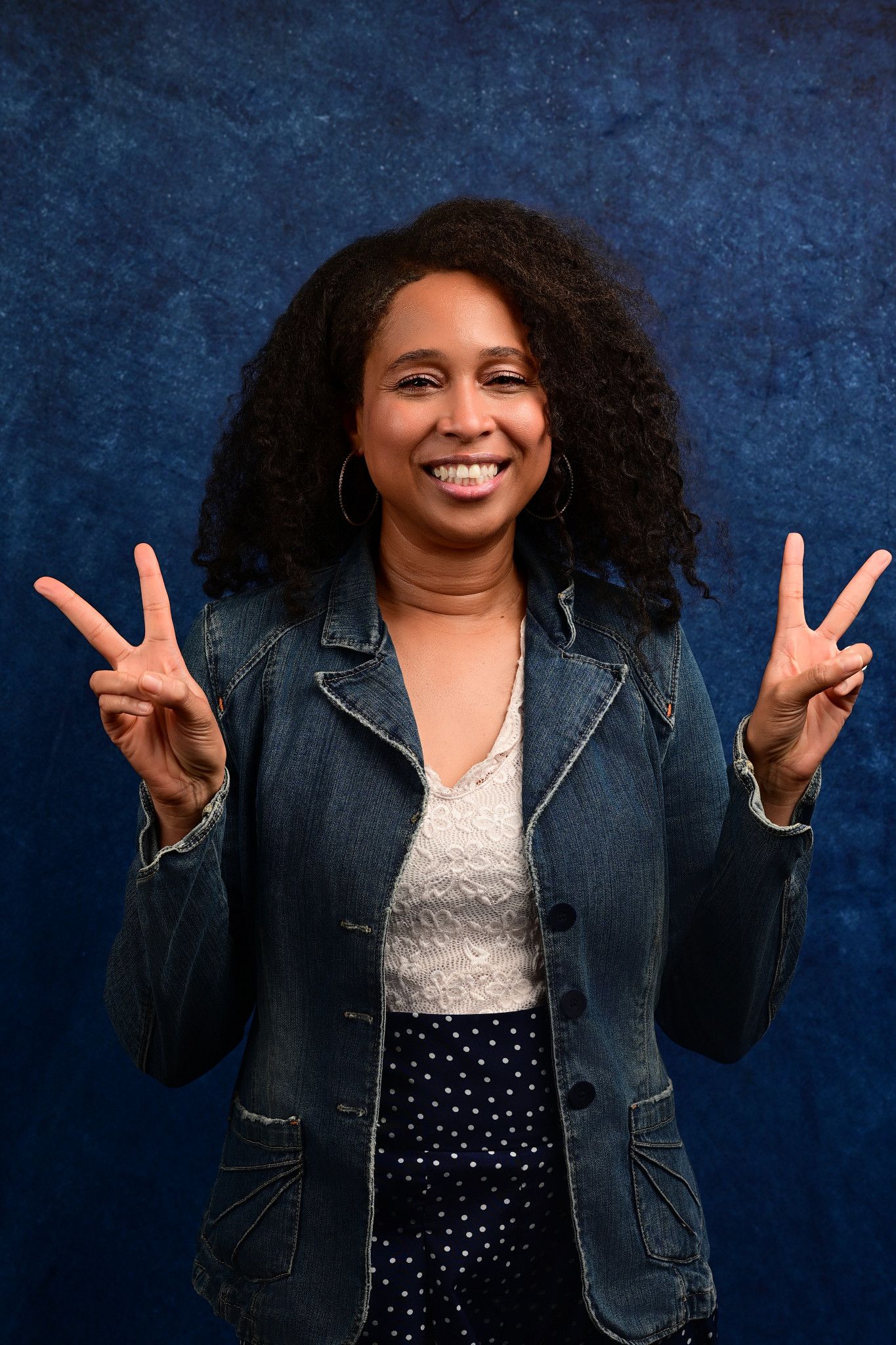 Smiling woman with curly dark hair flashes peace signs, wearing a denim jacket over a lace top and polka dot skirt against a blue backdrop.