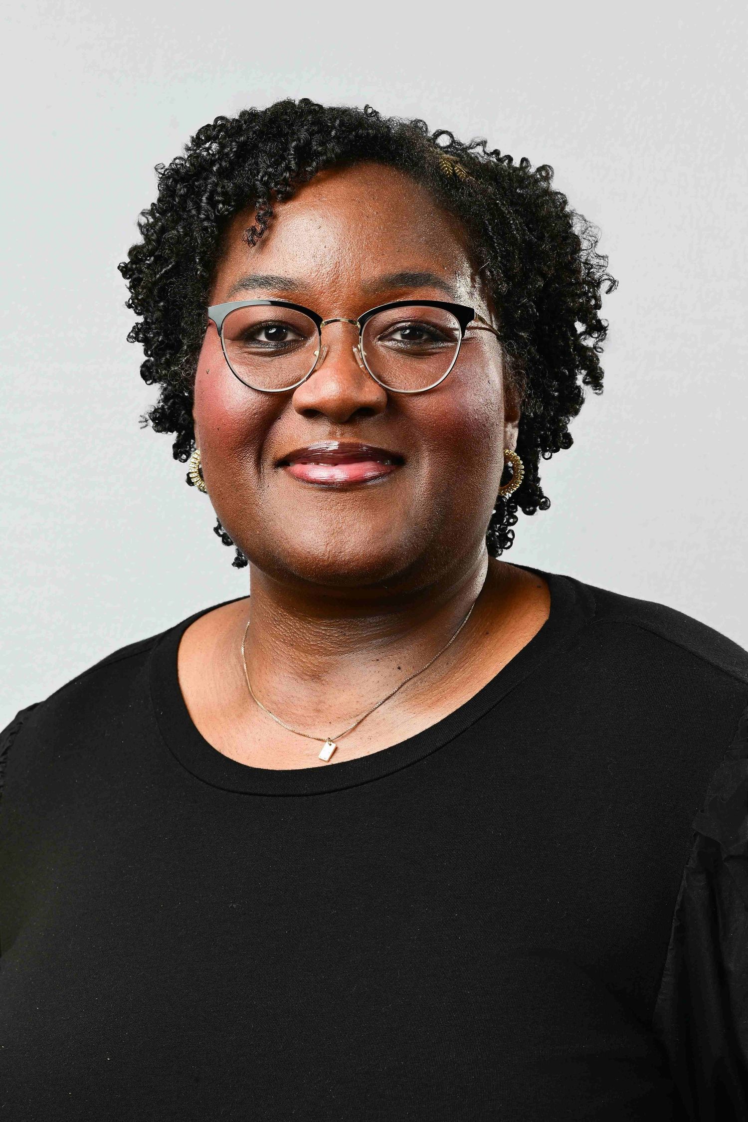 Smiling Black woman with curly hair and glasses wearing a black top and delicate necklace, professional headshot photo.