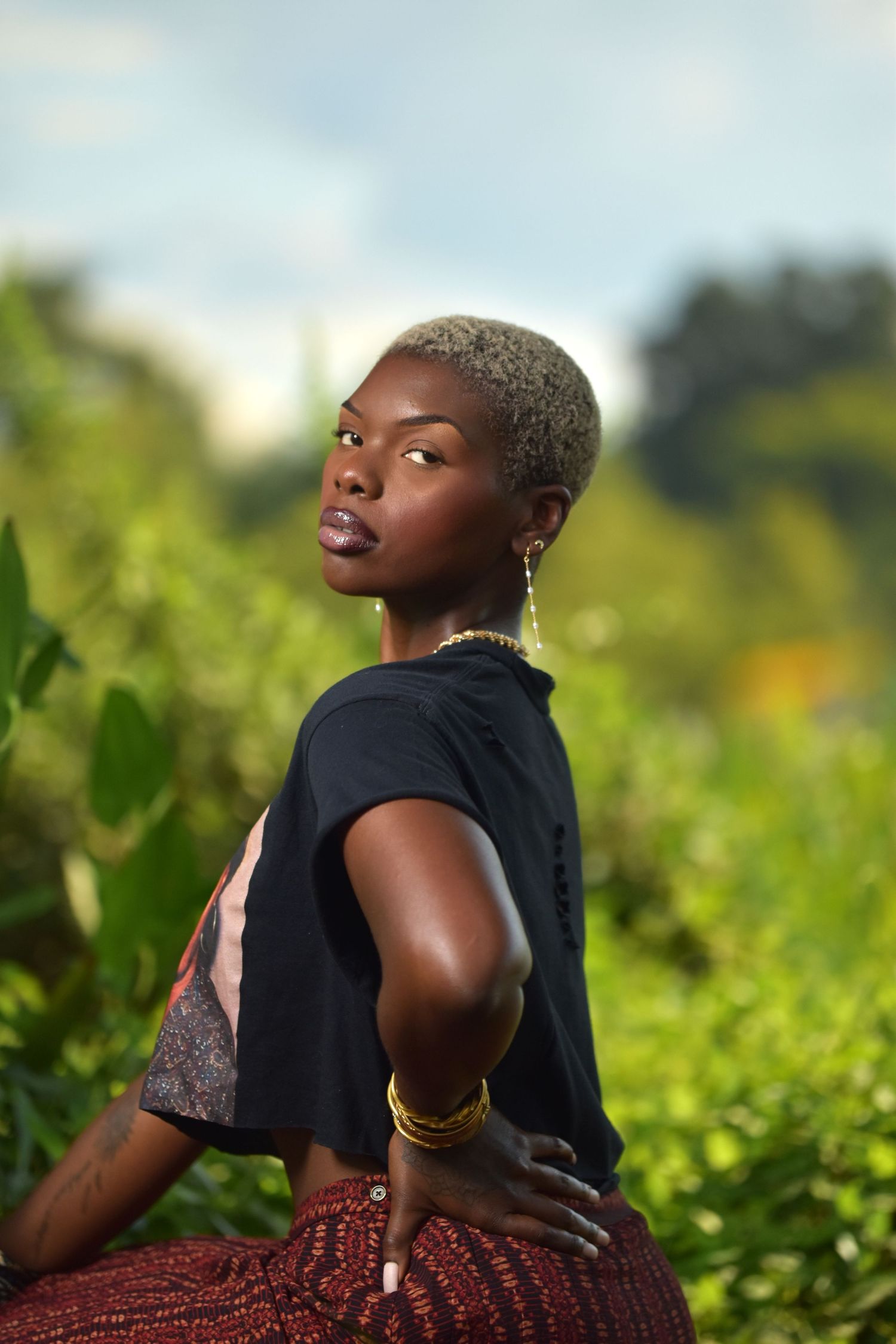 Stylish Black woman with short blonde hair posing outdoors in black tee and red patterned skirt with gold bangles.