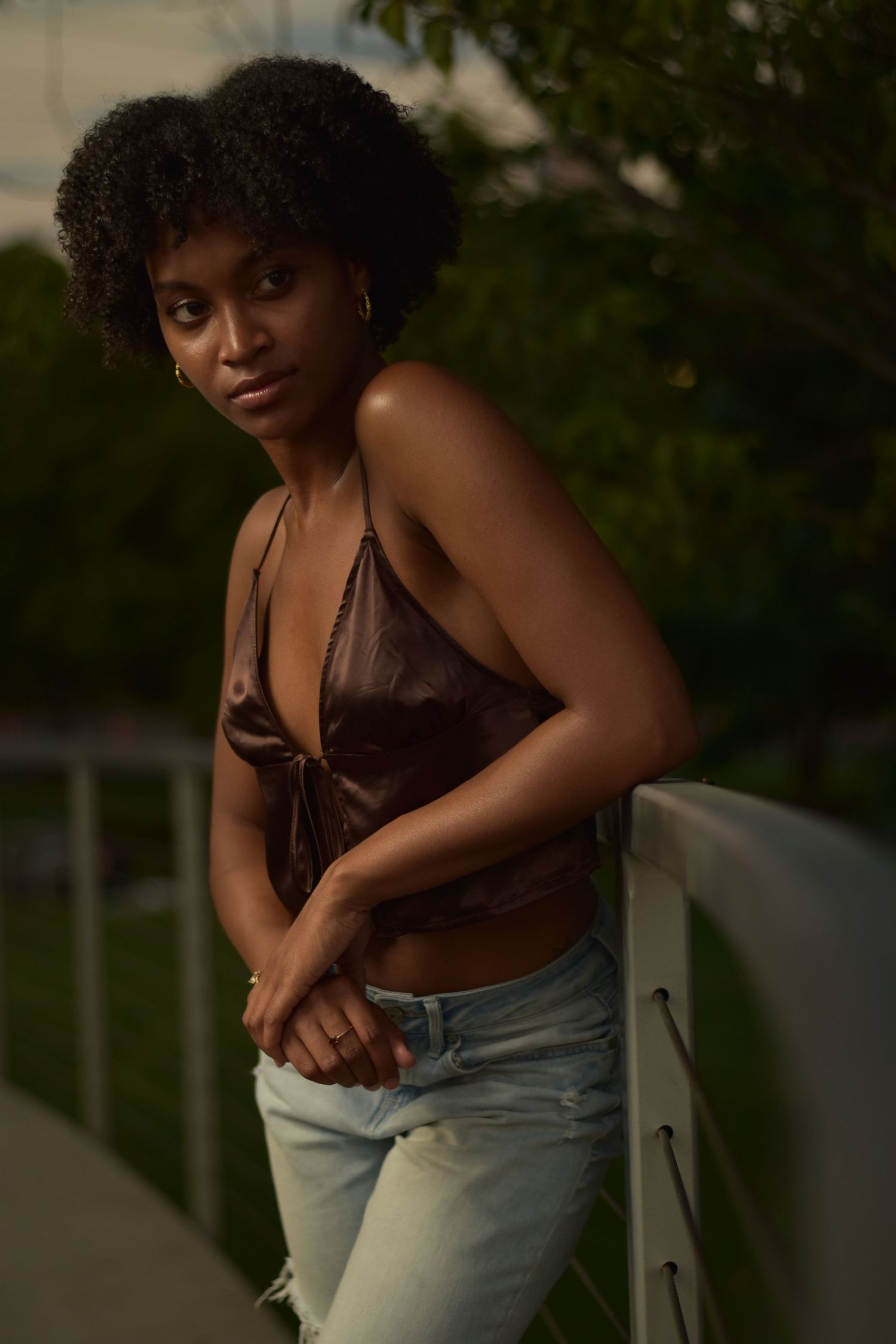 Young Black woman in brown halter top and ripped jeans leaning on white railing outdoors at dusk.