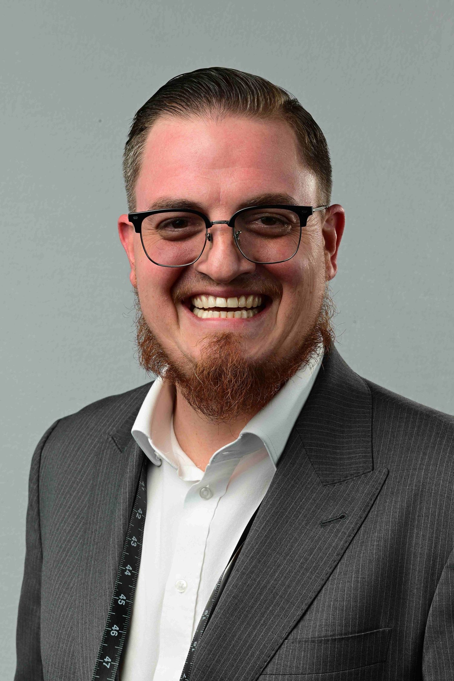 Smiling man with glasses and beard wearing a gray suit and white shirt for a professional headshot.