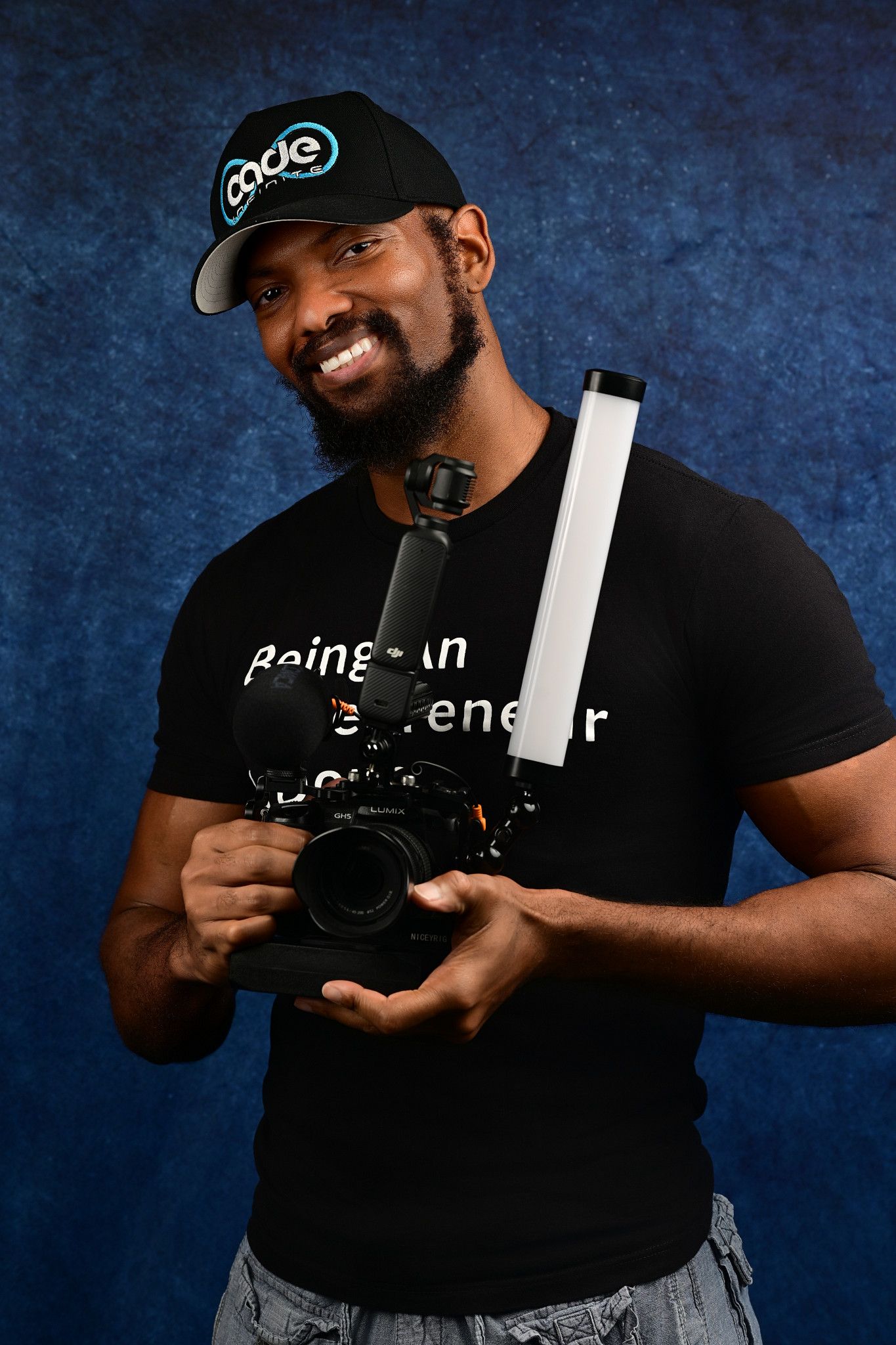 Smiling bearded man in a black cap and graphic tee holds a camera rig with a microphone and LED light wand against a blue backdrop.
