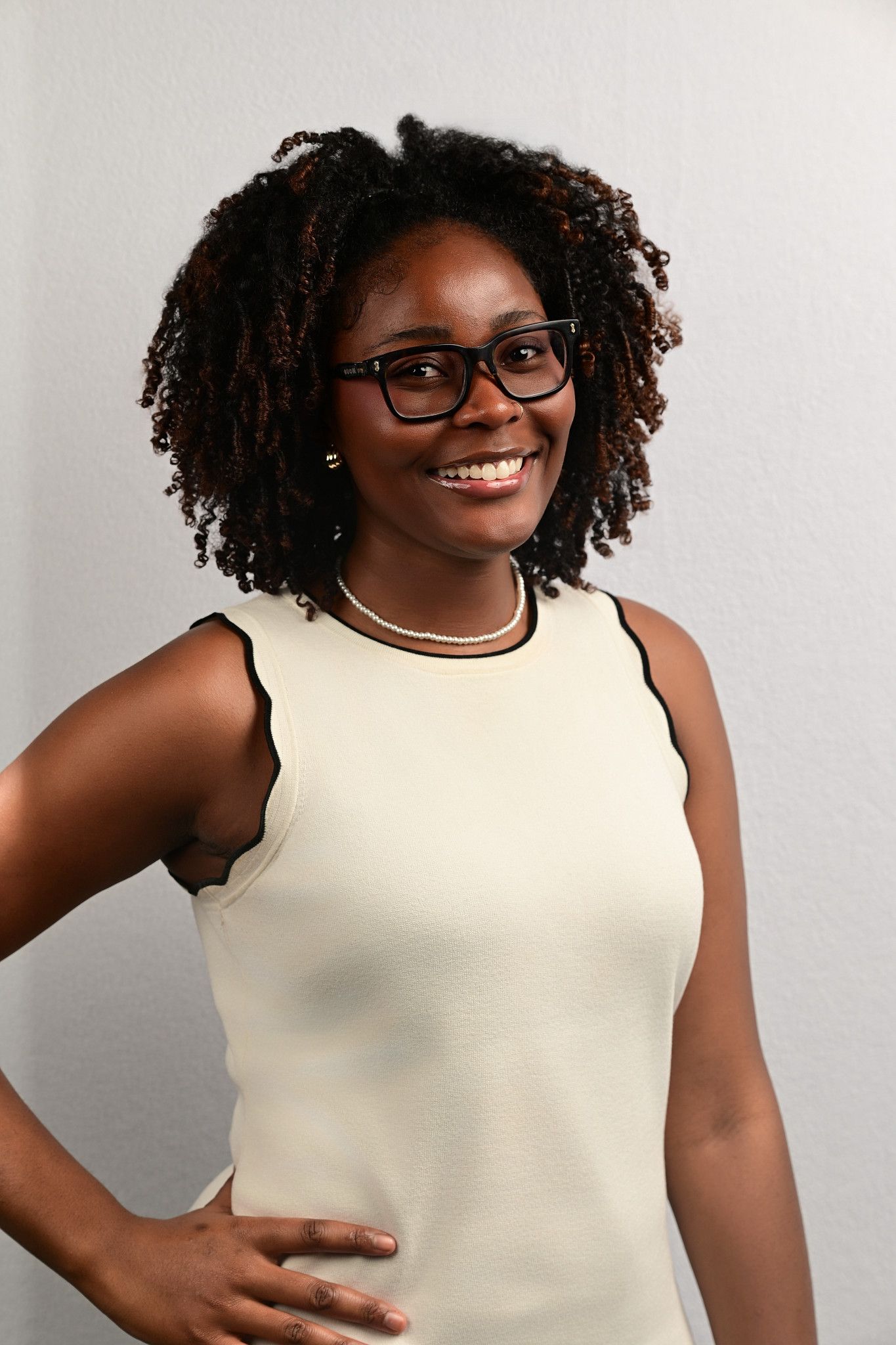 Smiling woman with curly hair and black-rimmed glasses poses with her hand on her hip in a cream sleeveless top and beaded necklace.