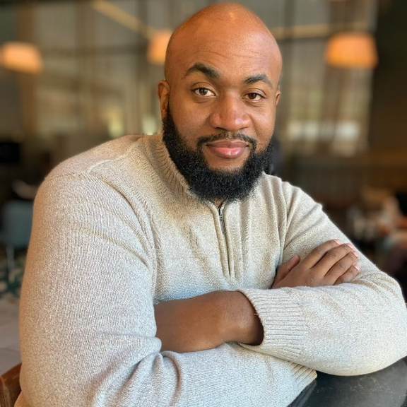 Professional Black man with beard in beige sweater sitting with arms crossed in modern restaurant setting.