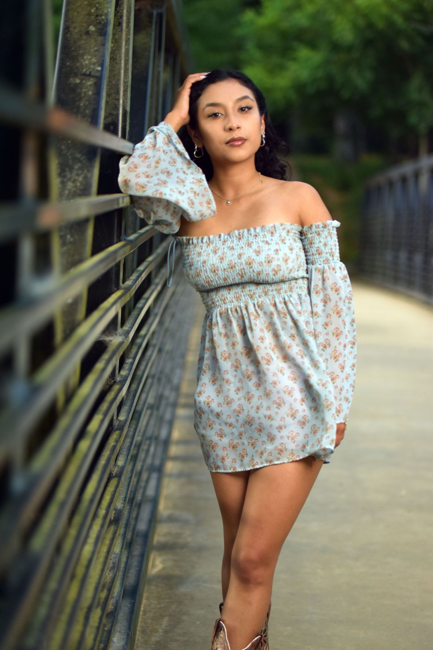 Young woman in floral off-shoulder mini dress poses gracefully on metal bridge railing.