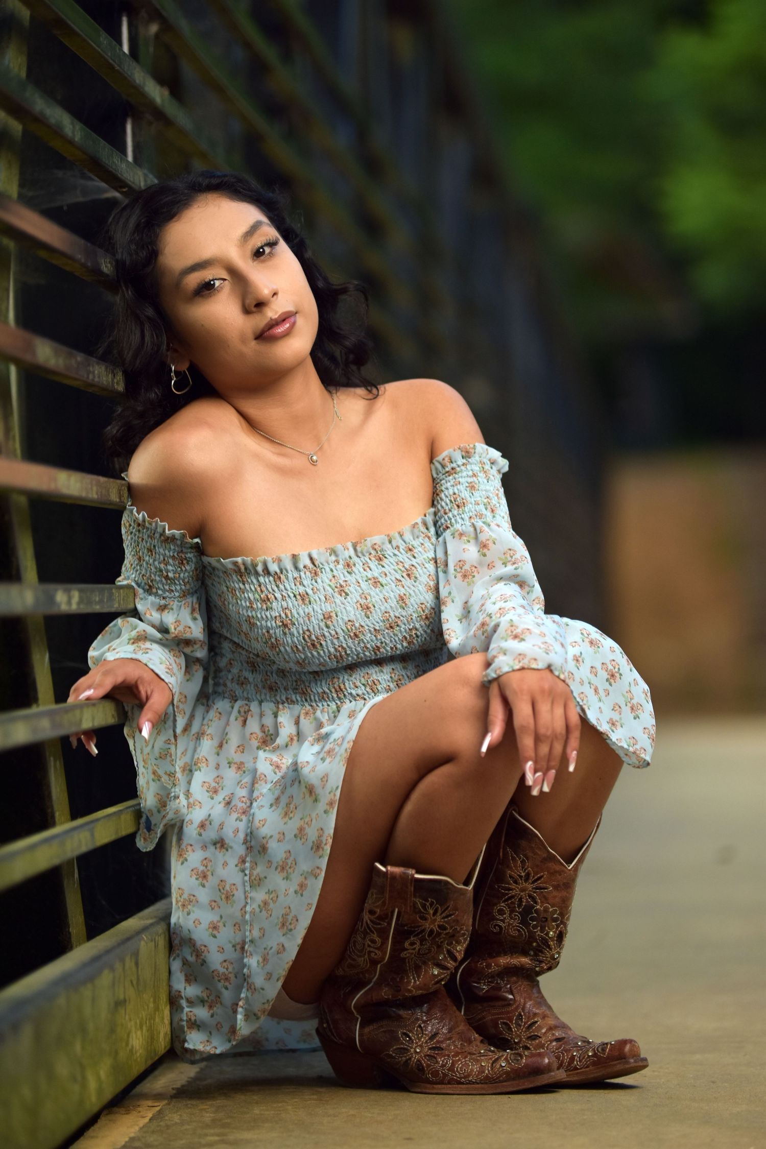 Young woman in light blue floral off-shoulder dress and cowboy boots sitting against metal fence.