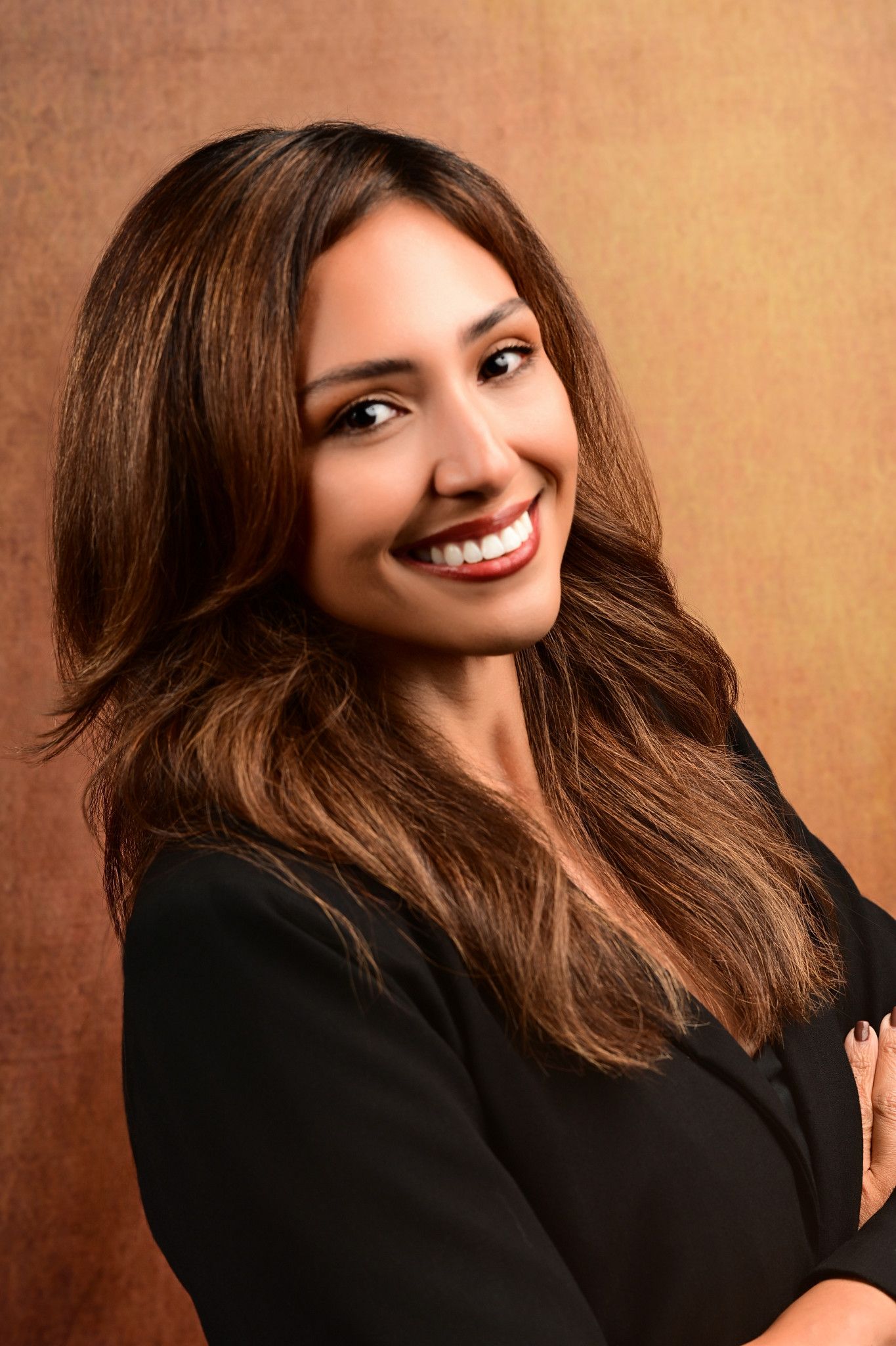Smiling woman with long layered brown hair and a black blazer poses with arms crossed against a warm bronze backdrop.