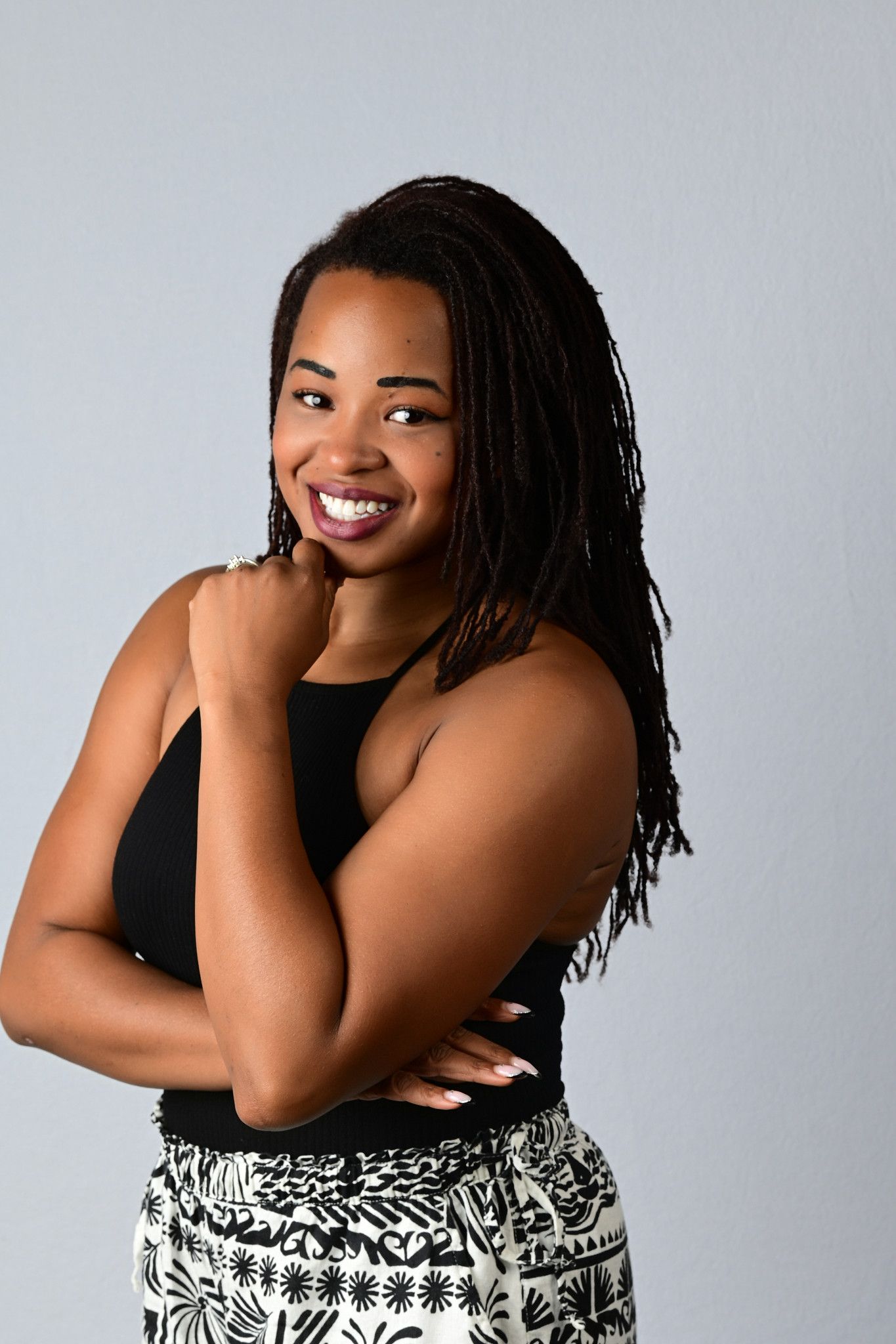 A smiling woman with long braids and a black top poses with her arms crossed and hand at her chin against a gray backdrop.