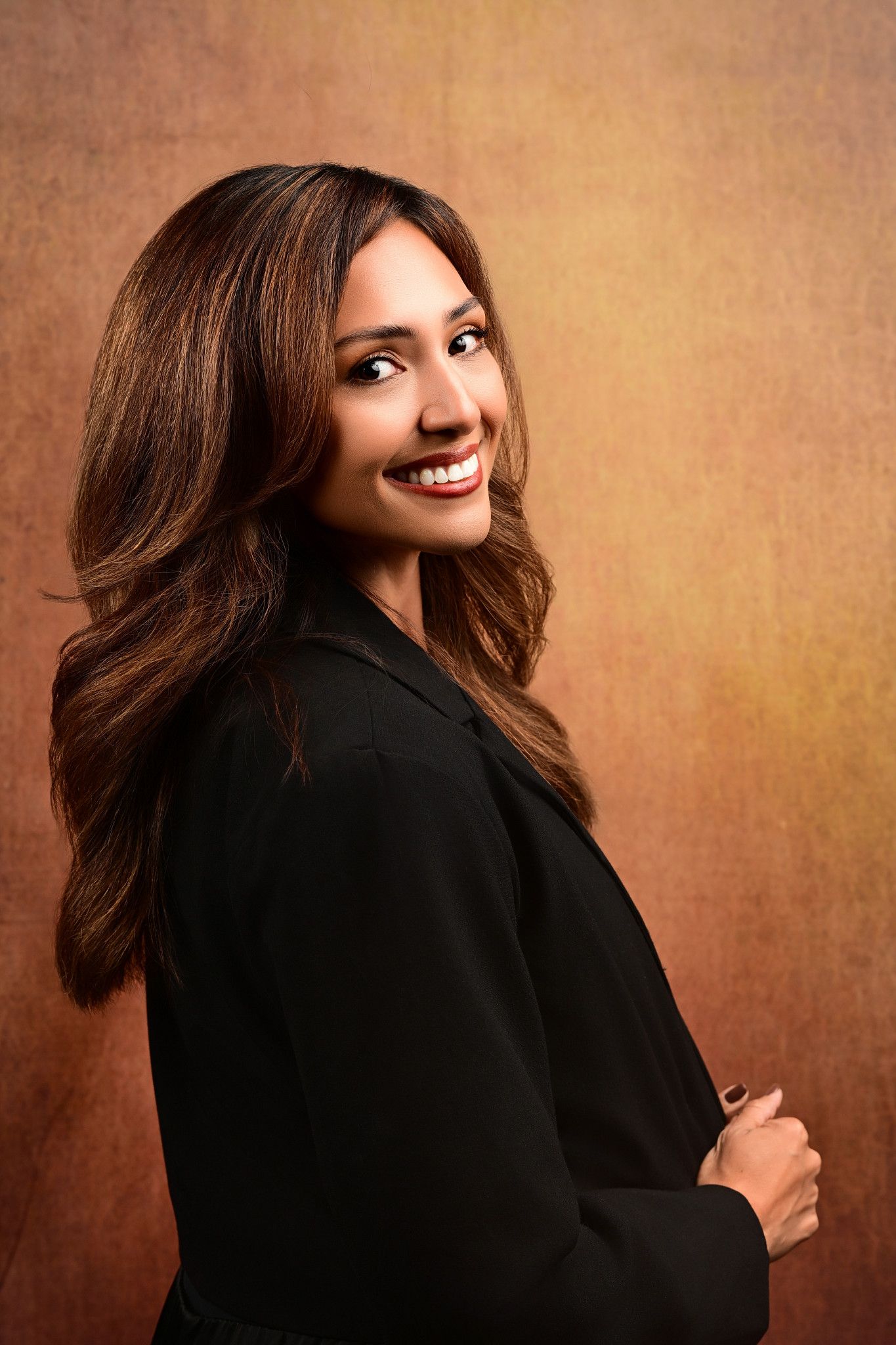 Smiling woman with long wavy brown hair in a black blazer poses over her shoulder against a warm golden-brown textured studio backdrop.