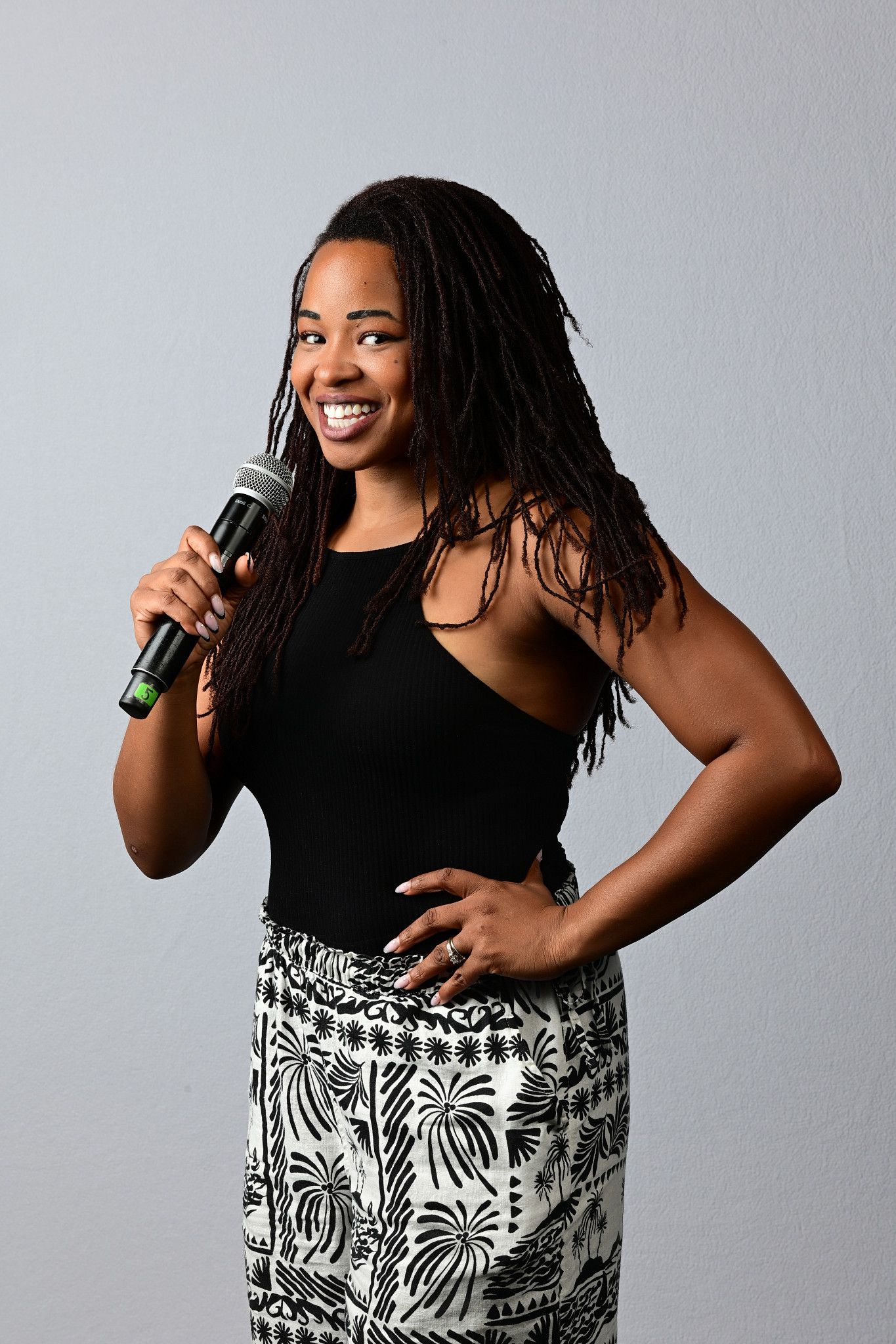 Smiling woman with long locs holds a microphone, wearing a black halter top and palm-print pants, standing with a hand on her hip.