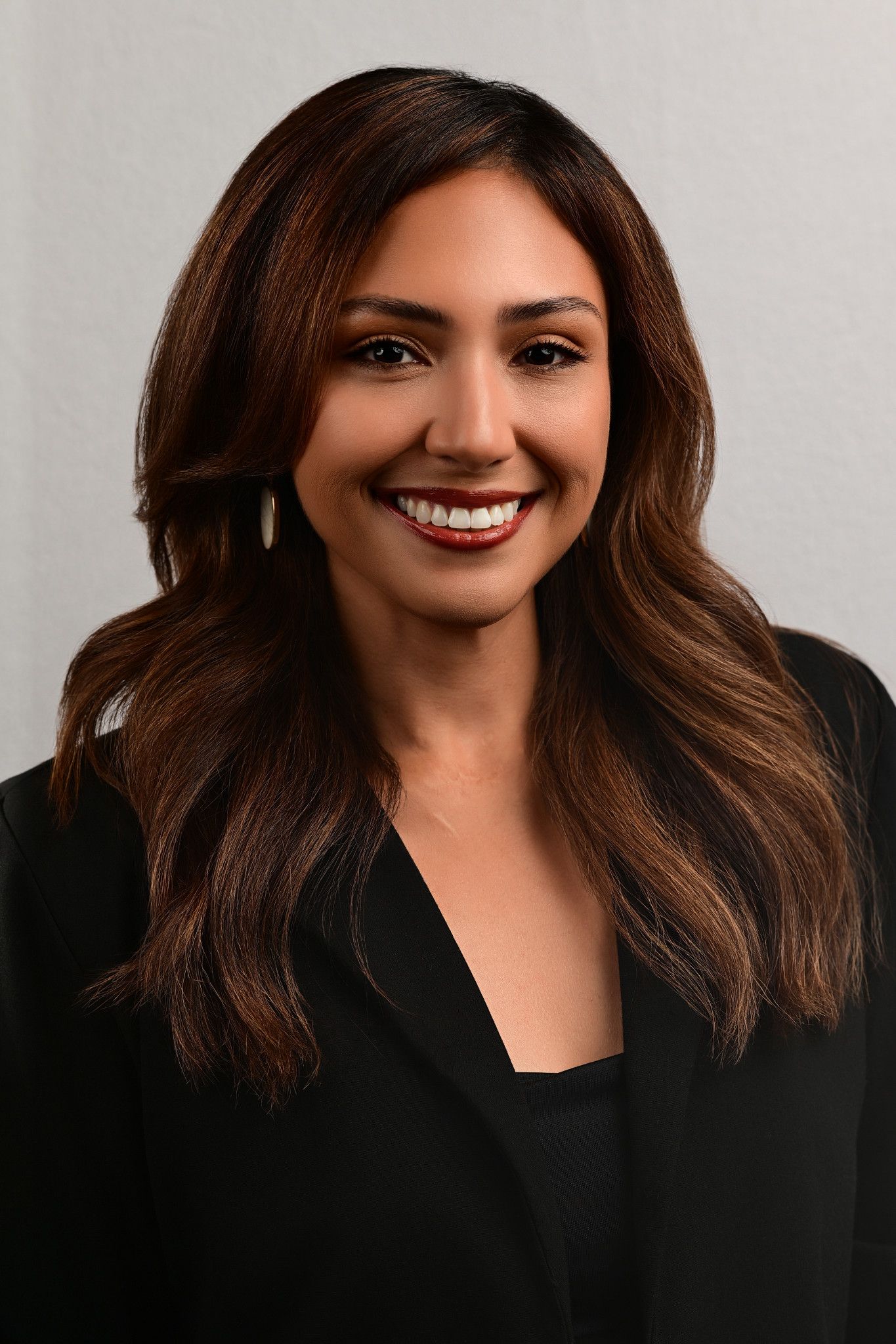 Smiling woman with long wavy brown hair and red lipstick wearing a black blazer in a professional headshot against a light gray background.