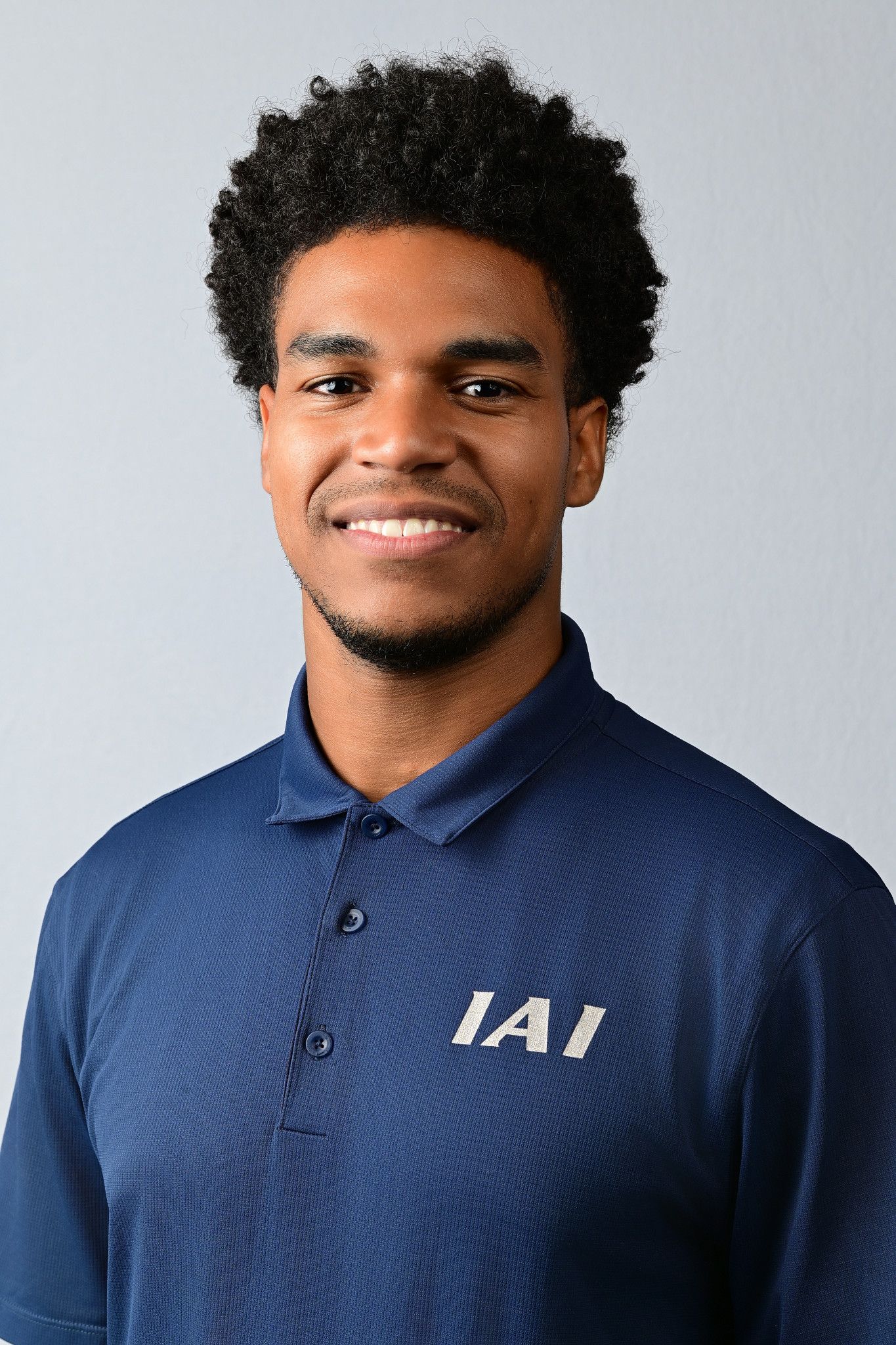 Young man smiling in a navy blue IAI polo shirt against a light gray background.