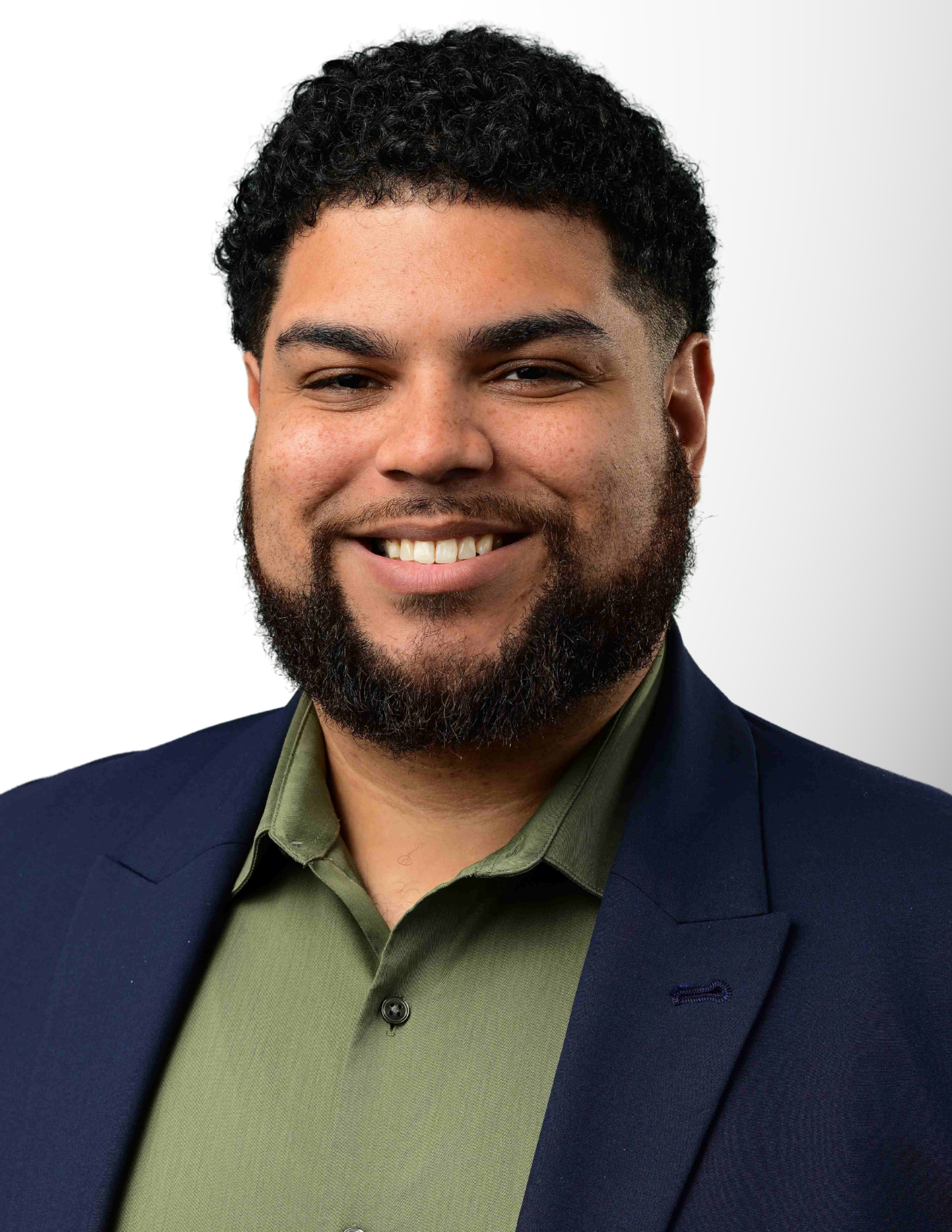 Smiling young man with beard wearing a green shirt and navy blazer against a white background.