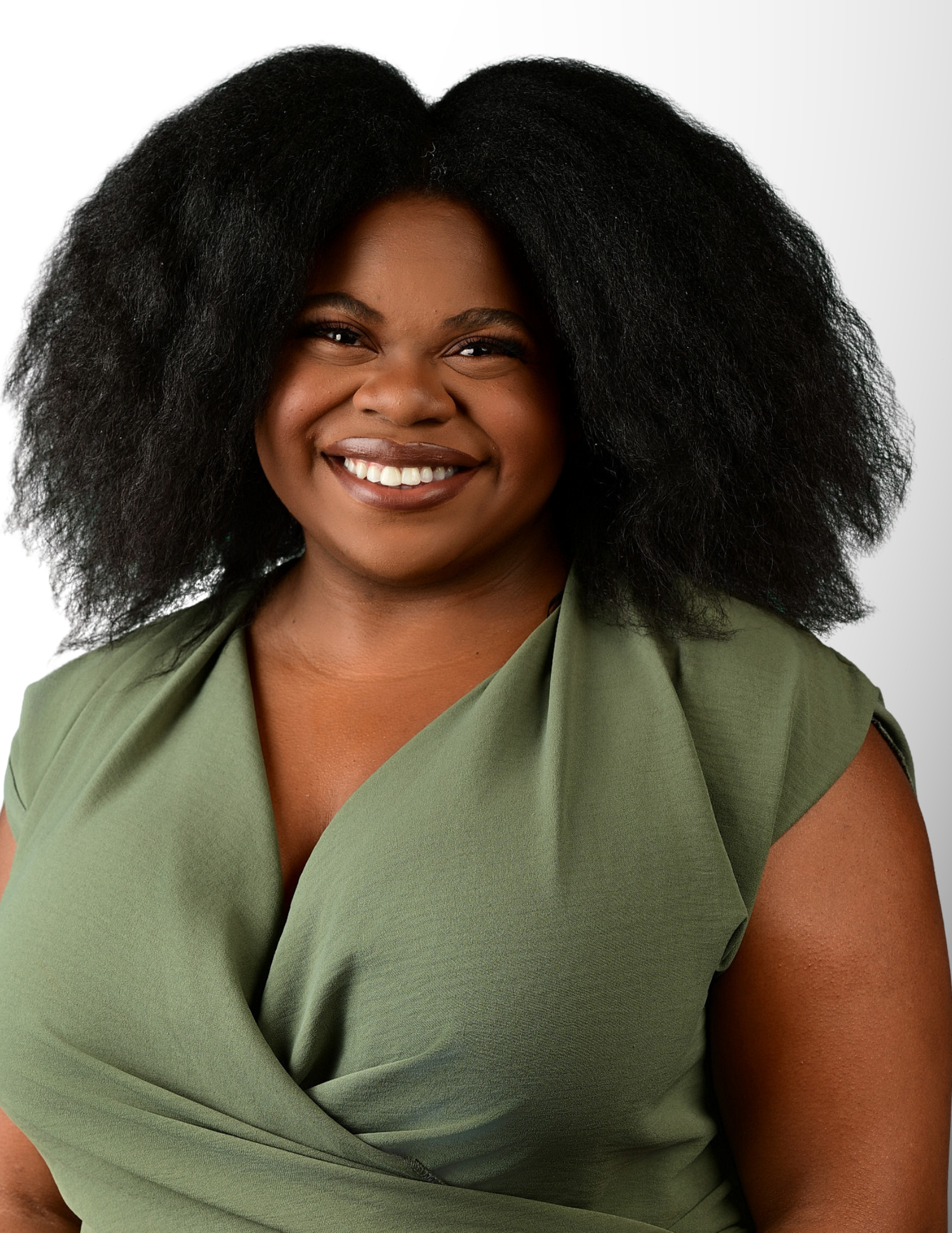 Smiling young Black woman with natural afro hair wearing a green wrap top against a white background.