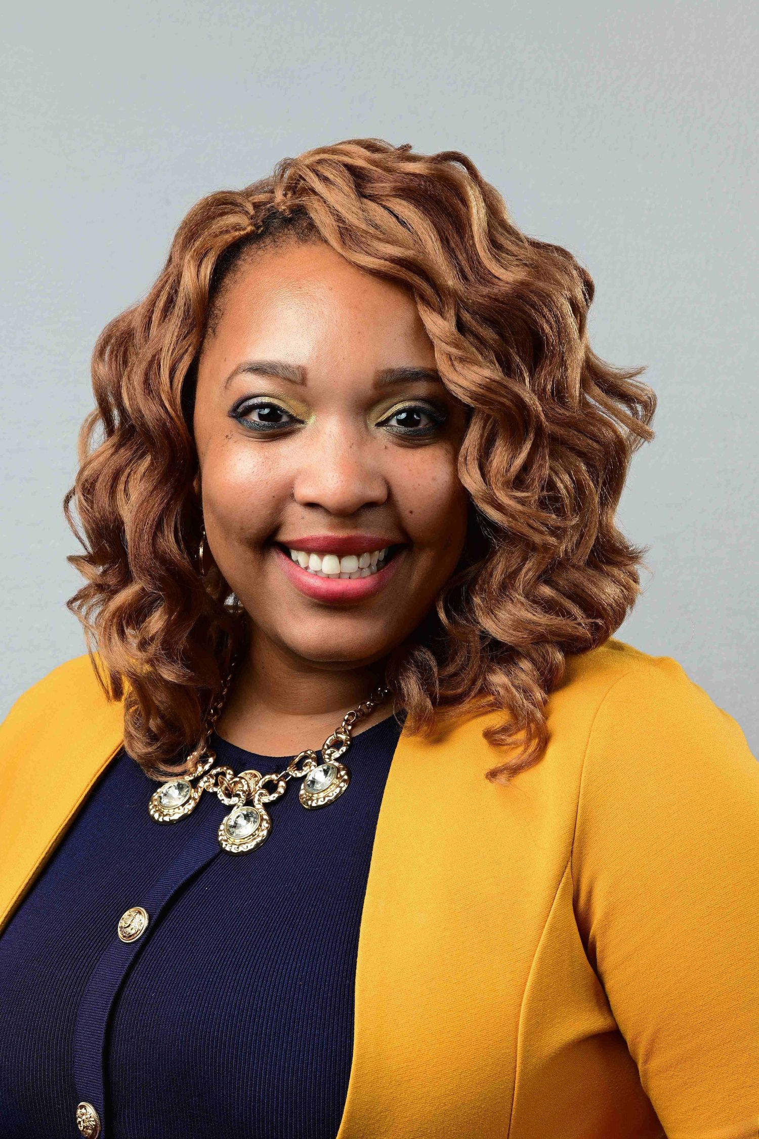 Professional headshot of a smiling woman with curly auburn hair wearing a yellow blazer and gold necklace.