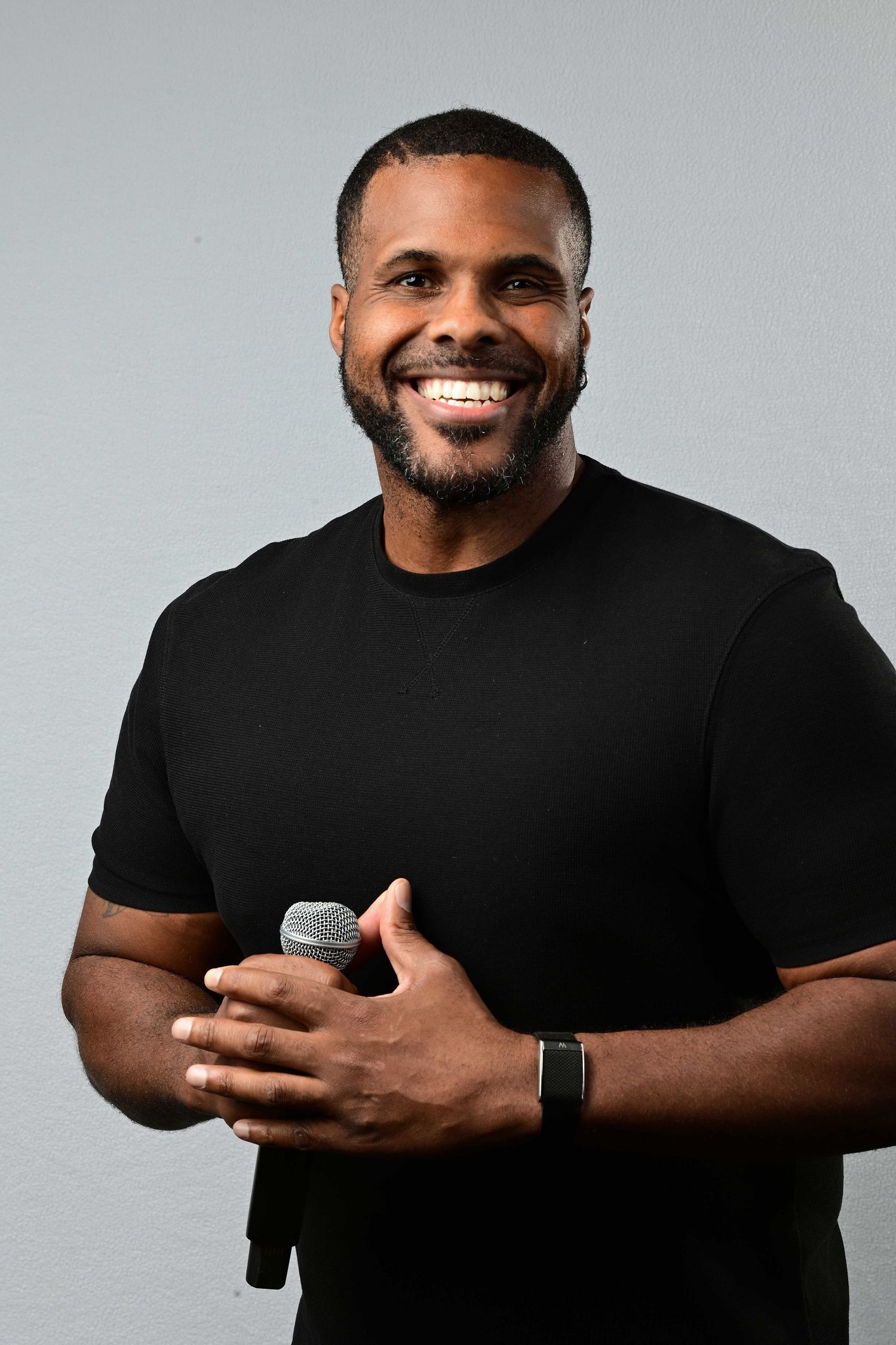 Smiling man with a short beard in a black t-shirt and smartwatch holds a microphone against a plain light gray background.