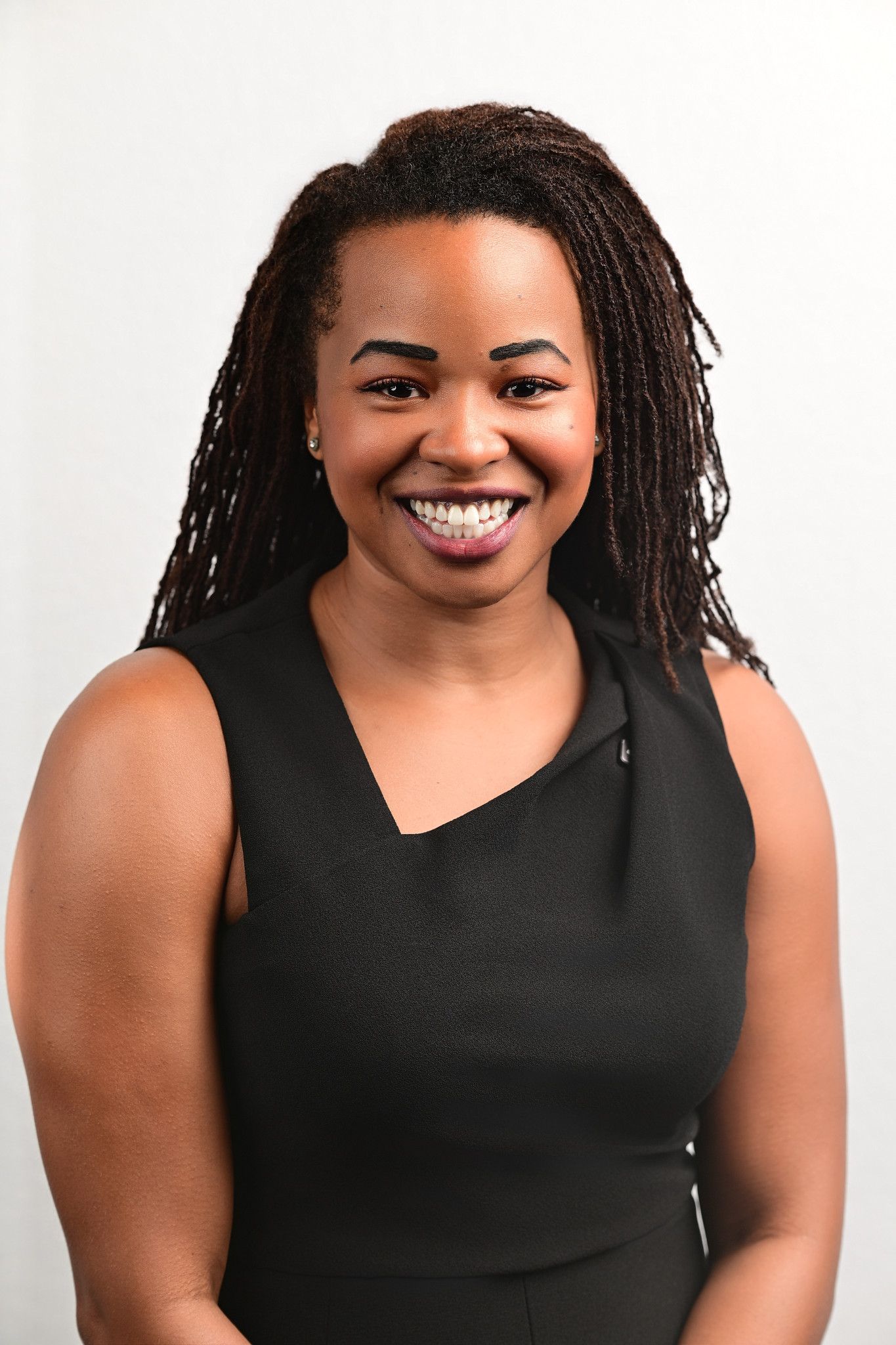 Smiling woman with long locs wearing a sleeveless black dress poses for a professional headshot against a plain light gray background.