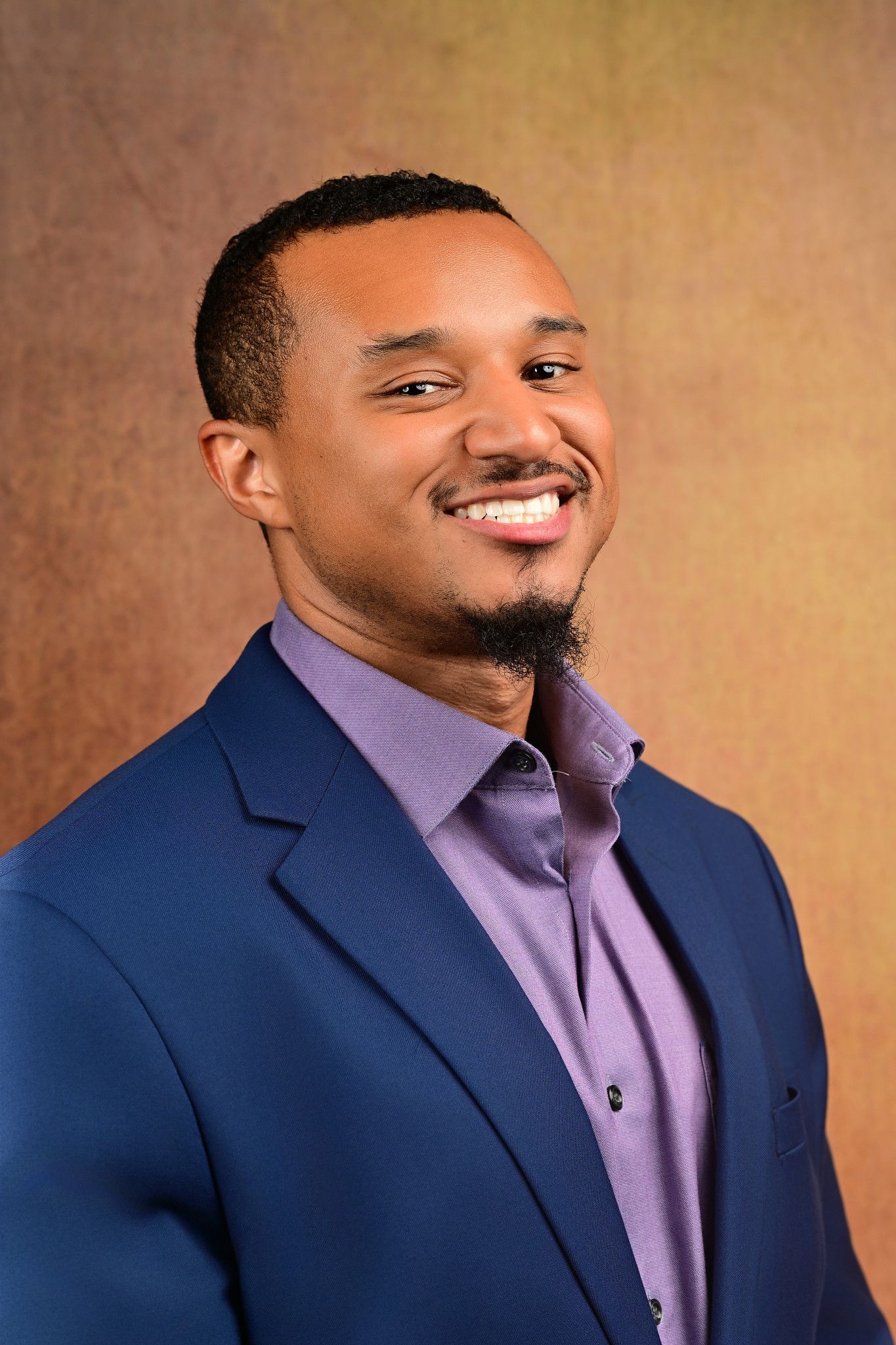 Smiling man with short hair and a goatee wears a blue blazer over a purple collared shirt against a warm brown textured backdrop.