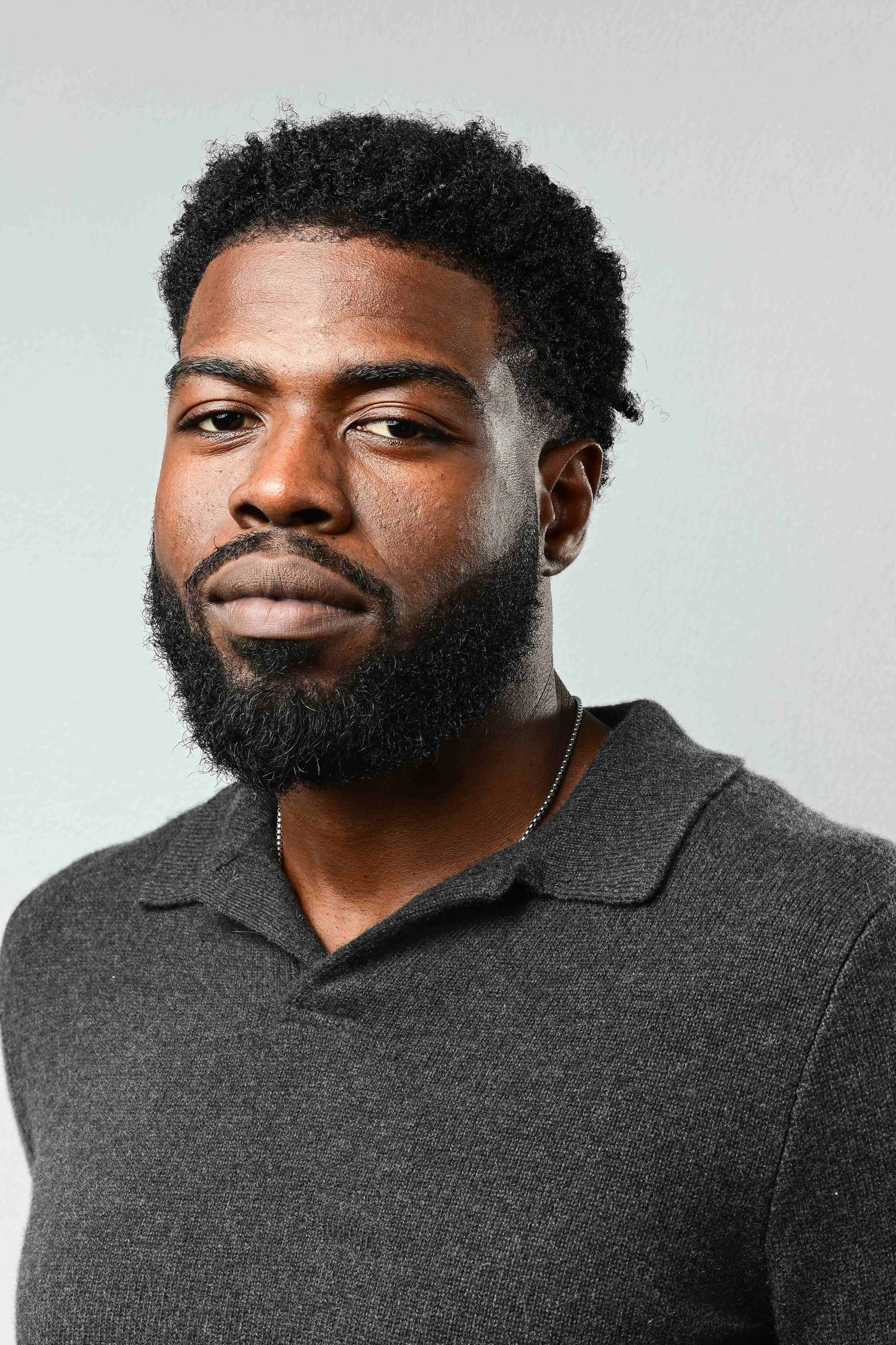 Young Black man with beard and natural hair wearing a dark gray polo shirt against a light gray background.