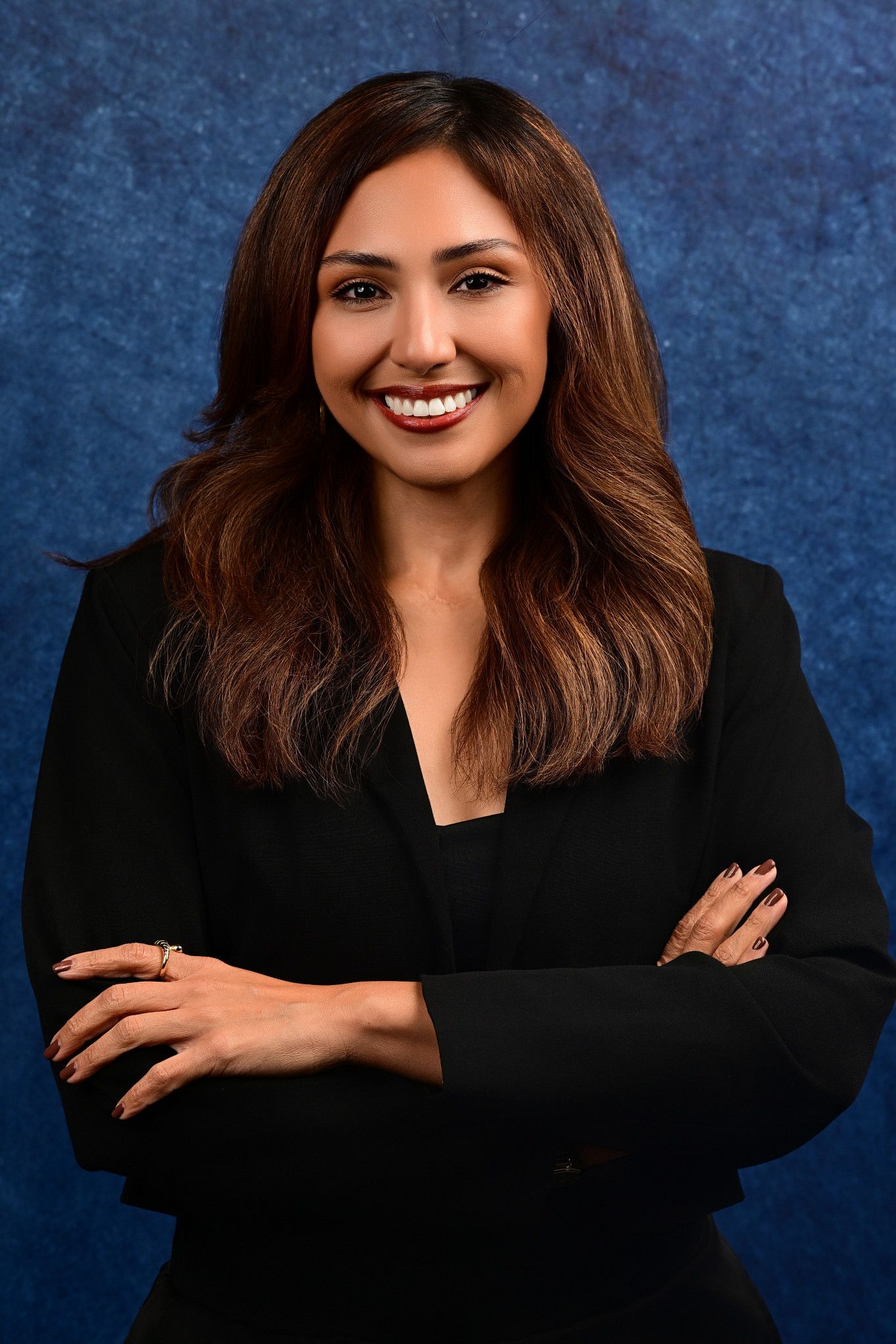 Smiling woman with long wavy brown hair wearing a black blazer poses with arms crossed against a blue textured studio backdrop.