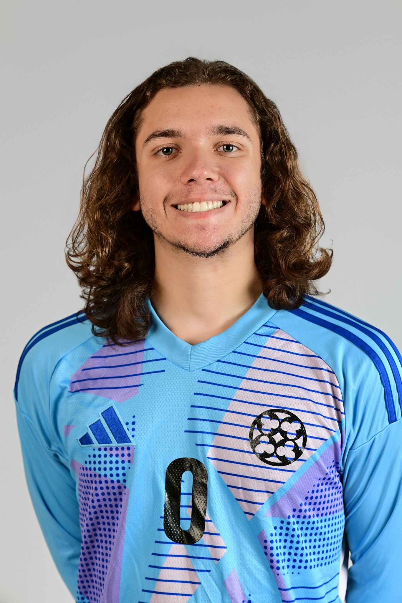 Male soccer goalkeeper smiling in light blue Adidas jersey with curly brown hair.